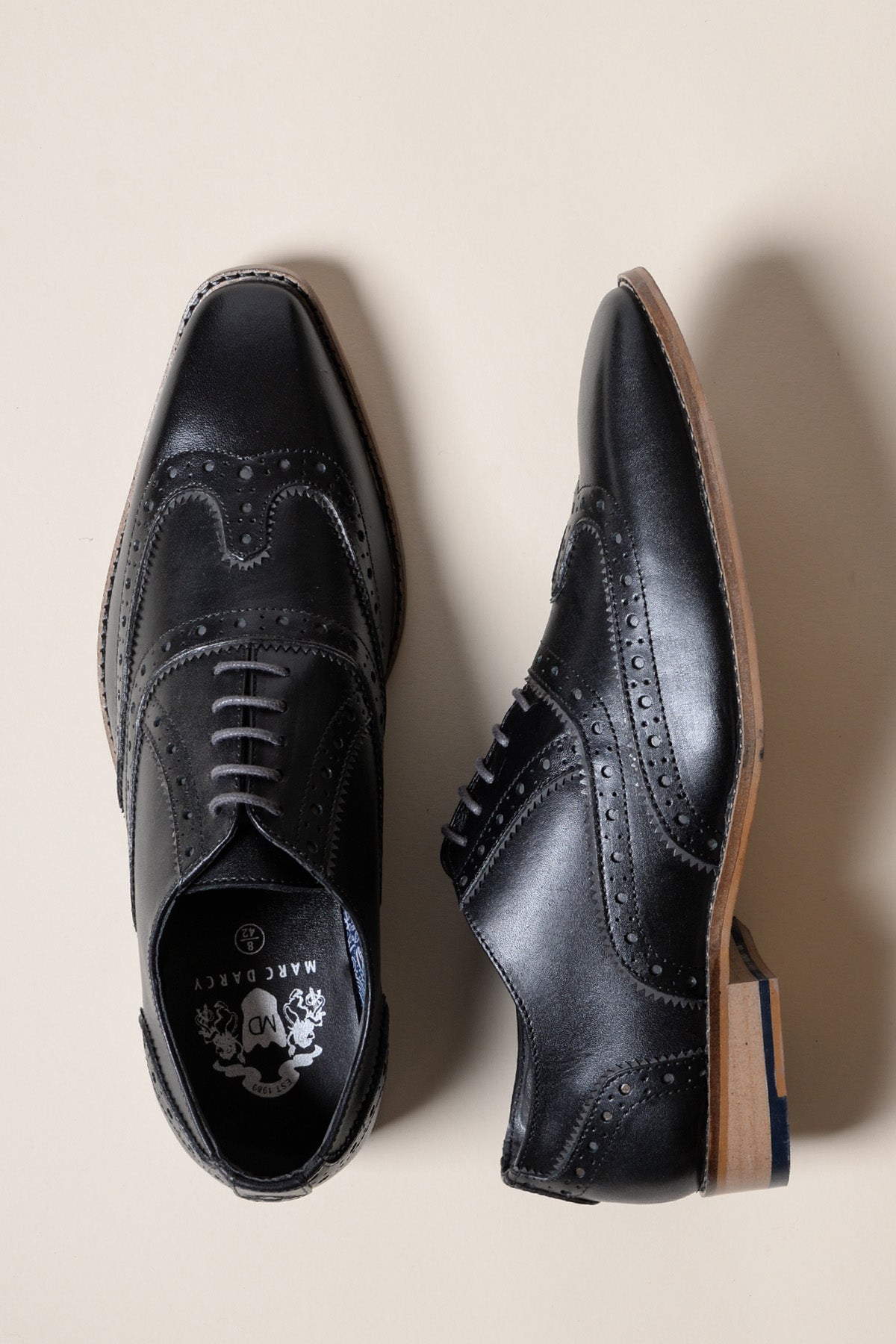 The MARCO Black Grey Leather Wingtip Oxford Shoe by Marc Darcy is shown on a light beige background; one shoe is seen from above and the other from the side, emphasizing its decorative perforations, sole, and heel.