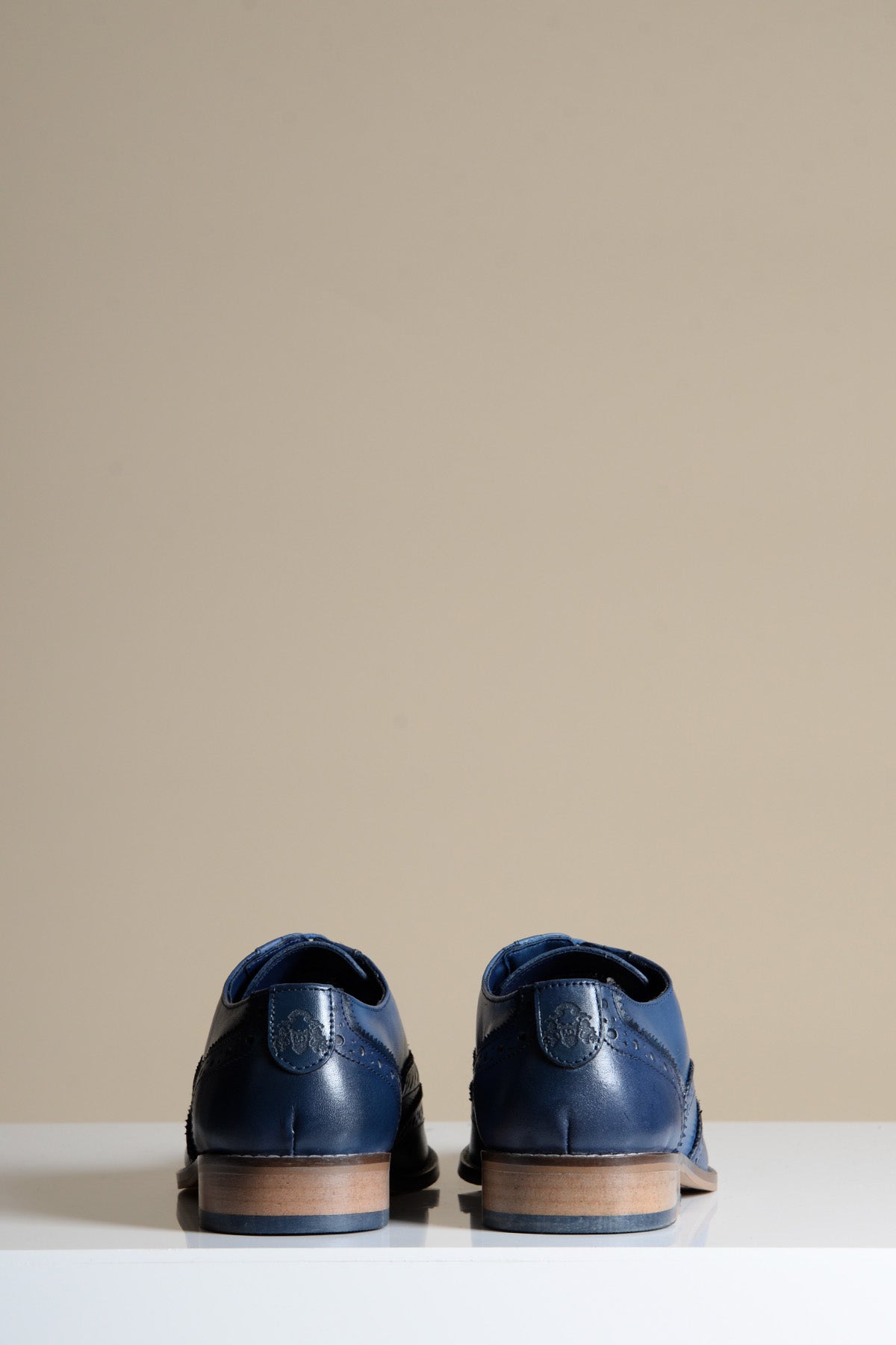 Rear view of two MARCO - Navy Blue Leather Wingtip Oxford Shoes by Marc Darcy, featuring full grain leather and brown soles, placed side-by-side on a white surface with a beige background.