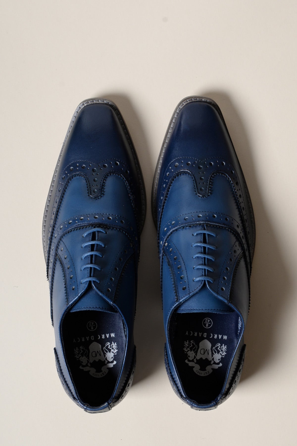 The MARCO - Navy Blue Leather Wingtip Oxford Shoe by Marc Darcy features shiny full grain leather and hand-stitched brogue detailing, displayed facing upward on a light beige background.