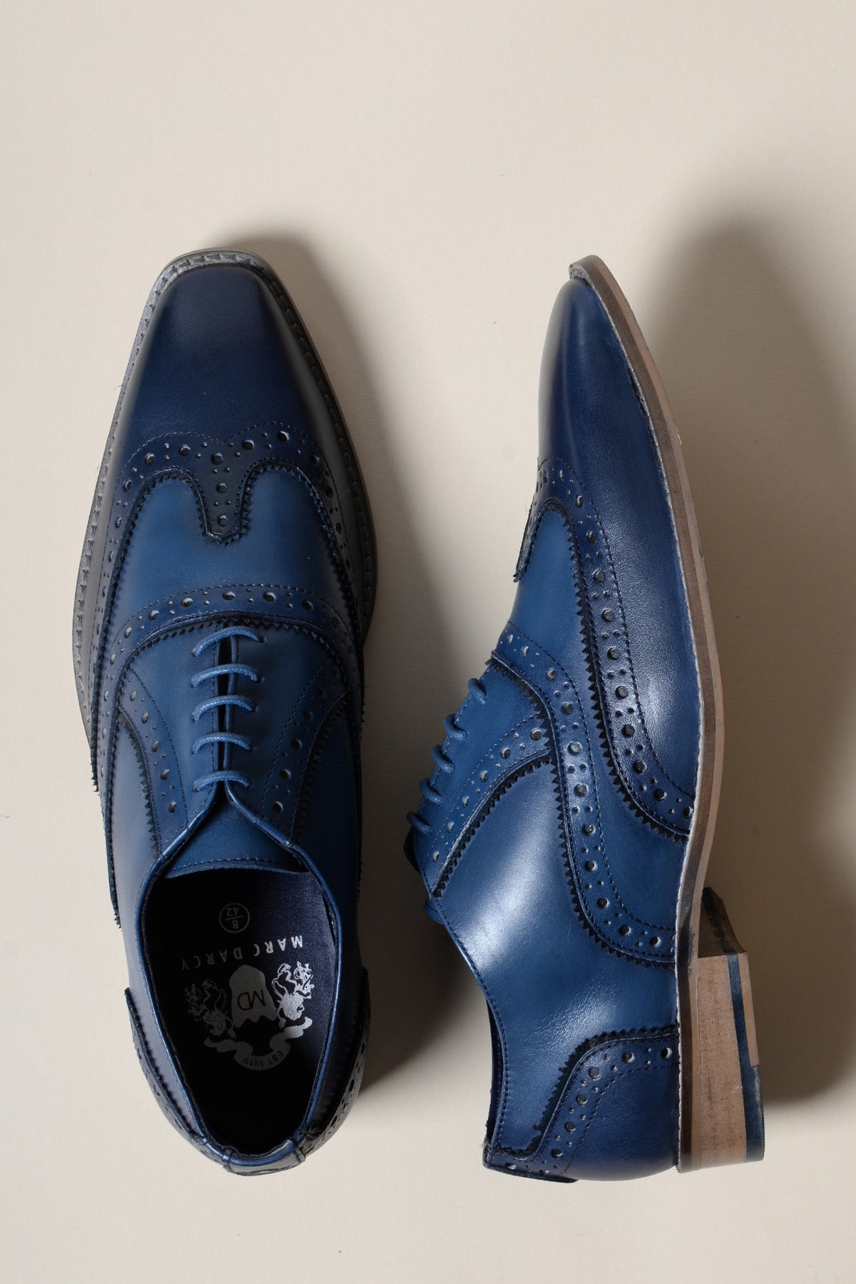 The MARCO Navy Blue Leather Wingtip Oxford Shoe by Marc Darcy is crafted from full grain leather with hand-stitched detailing and decorative perforations, shown with one shoe flat and the other on its side against a light beige background.