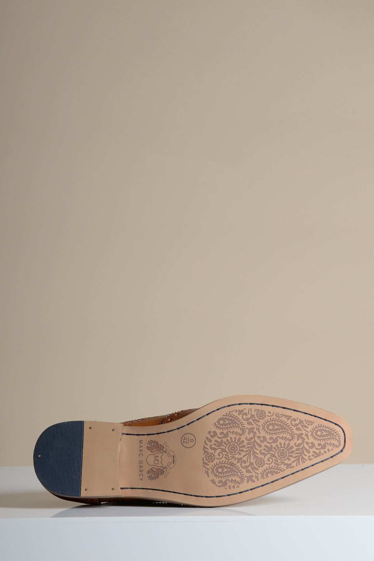 A close-up, angled shot of the sole of the MARCO - Tan Navy Leather Wingtip Oxford Shoe by Marc Darcy. The brown leather sole features intricate paisley detailing and displays the Marc Darcy logo.