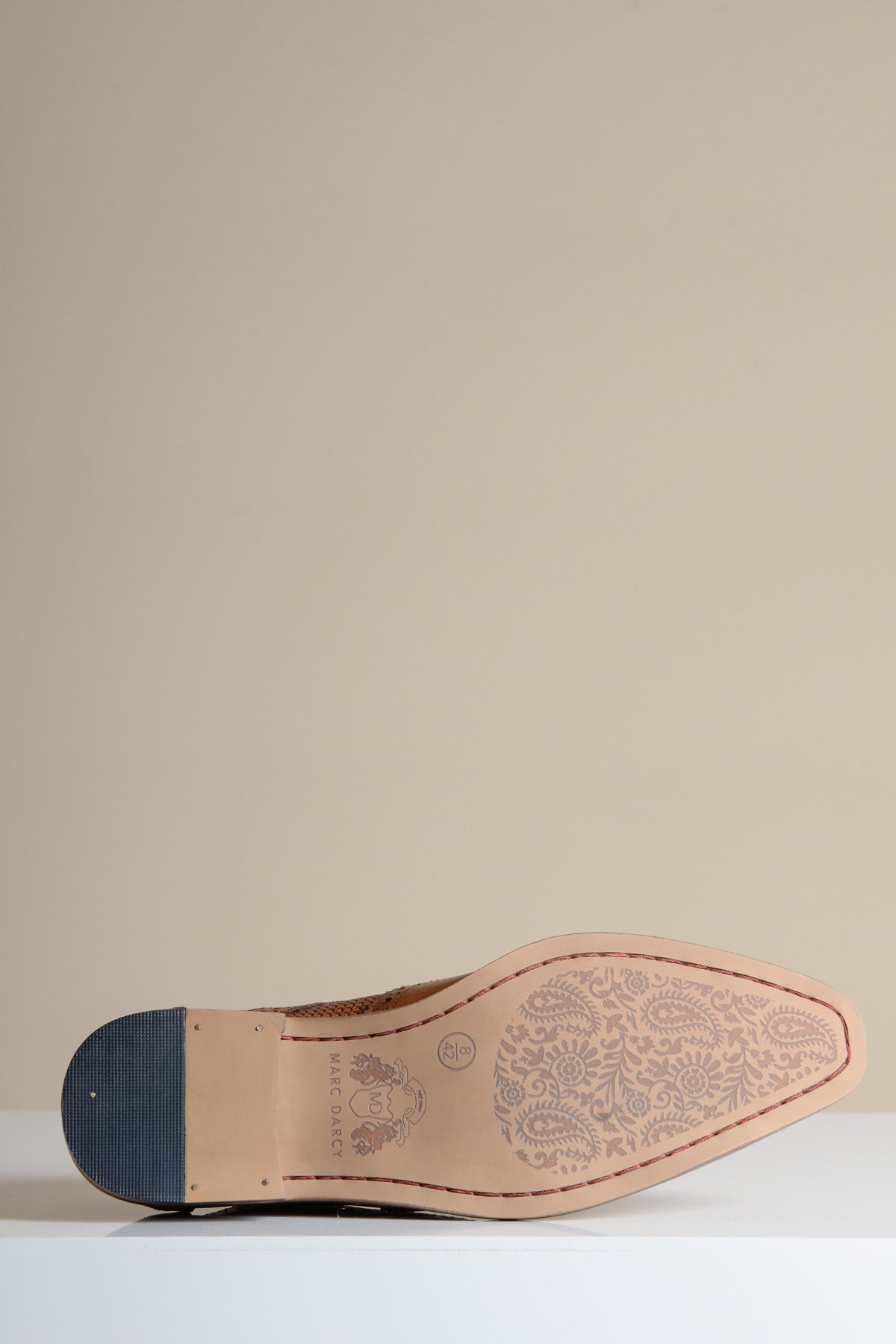 A close-up of the sole of the marcdarcy BRANDON Dark Tan Leather Wingtip Brogue Shoe, showing an intricate floral pattern and logo near the heel, set against a neutral beige background.