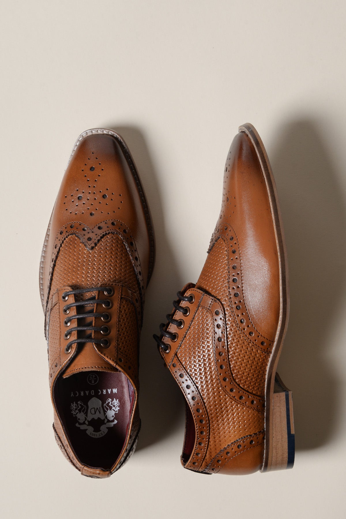 The marcdarcy BRANDON is a dark tan leather wingtip brogue shoe with hand-stitched full grain leather, decorative perforations, and textured detailing. Shown on a neutral background, one shoe faces up and the other is on its side.