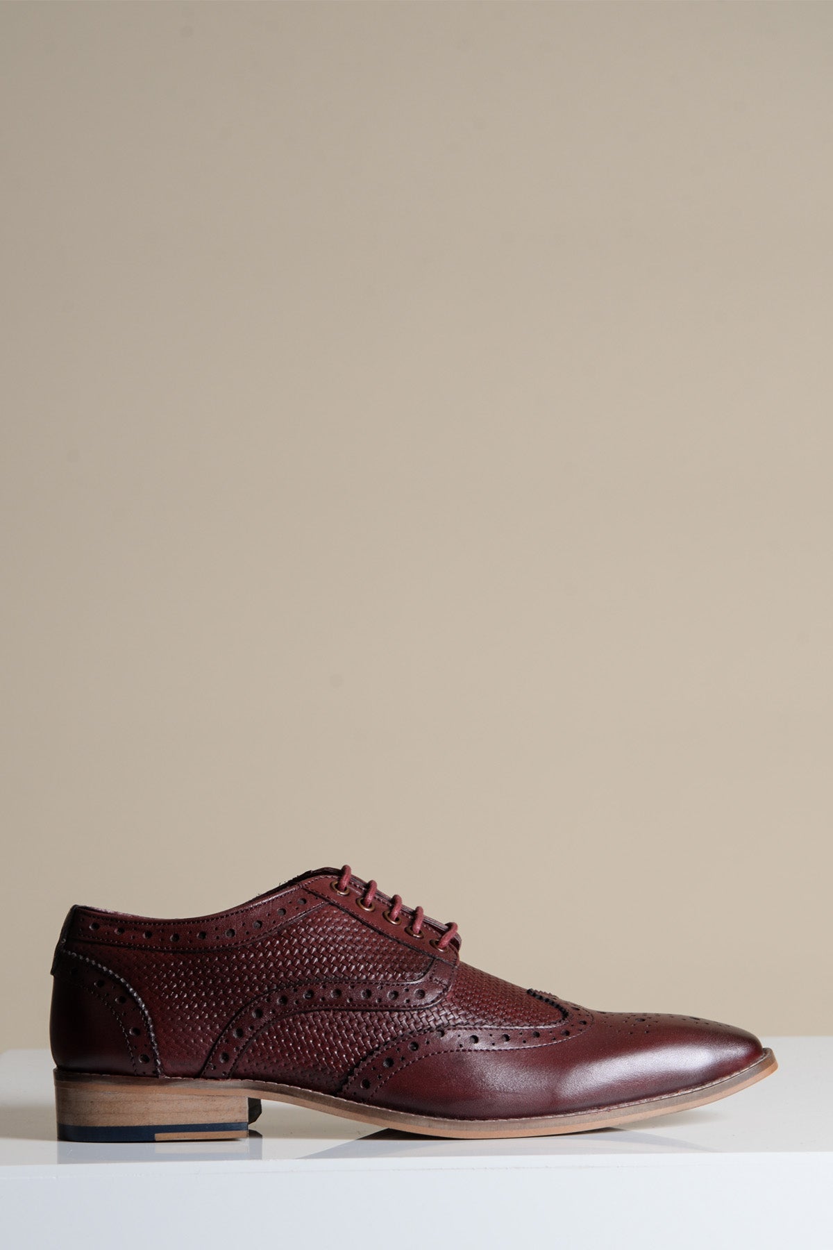 The BRANDON Bordeaux Burgundy Leather Wingtip Brogue Shoe by marcdarcy is shown in profile on a white surface with a beige background, highlighting its decorative perforations and signature style for comfortable men's footwear.