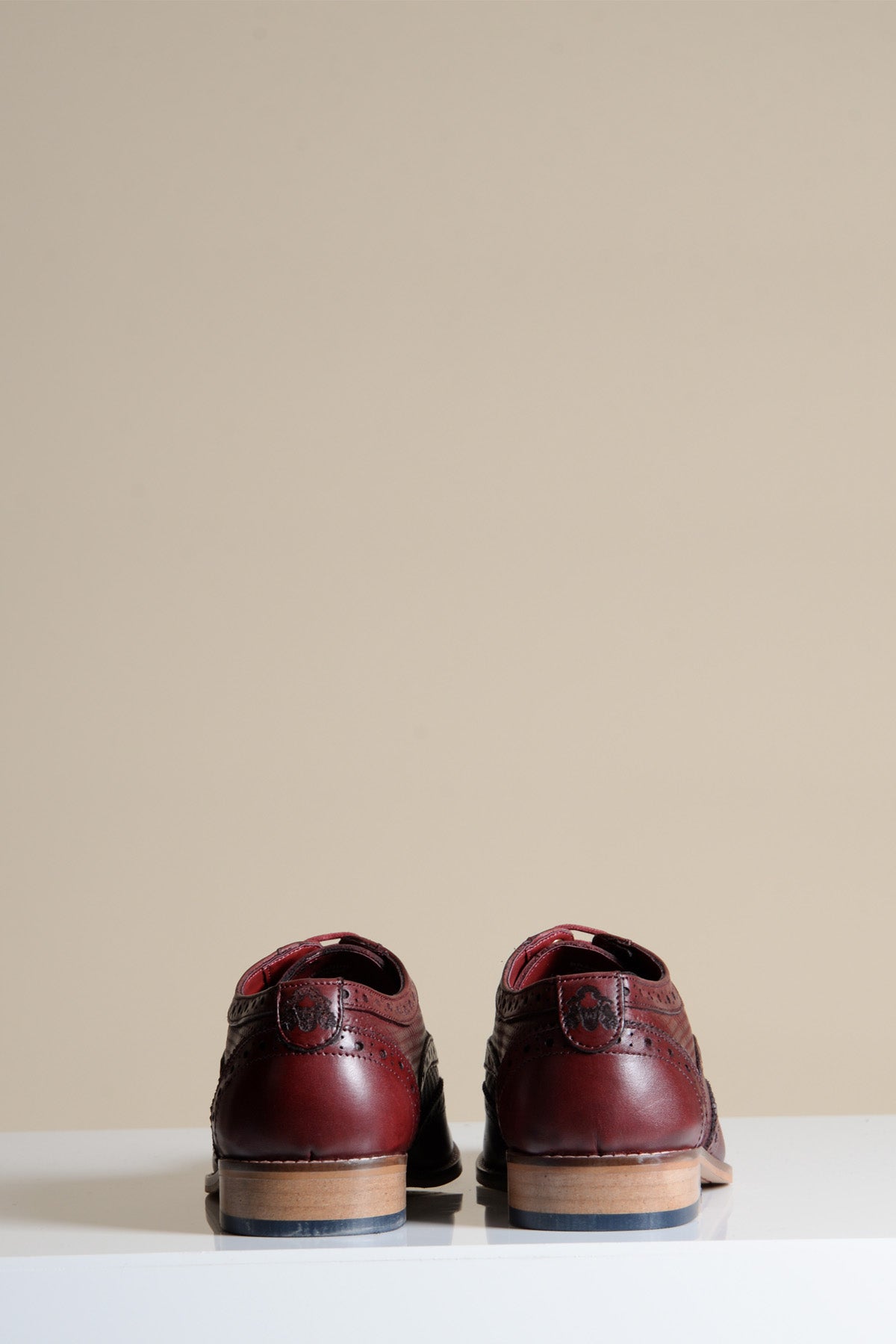 The BRANDON - Bordeaux Burgundy Leather Wingtip Brogue Shoe by marcdarcy rests on a white surface against a beige background, showcasing stylish and durable men's footwear crafted for comfort.