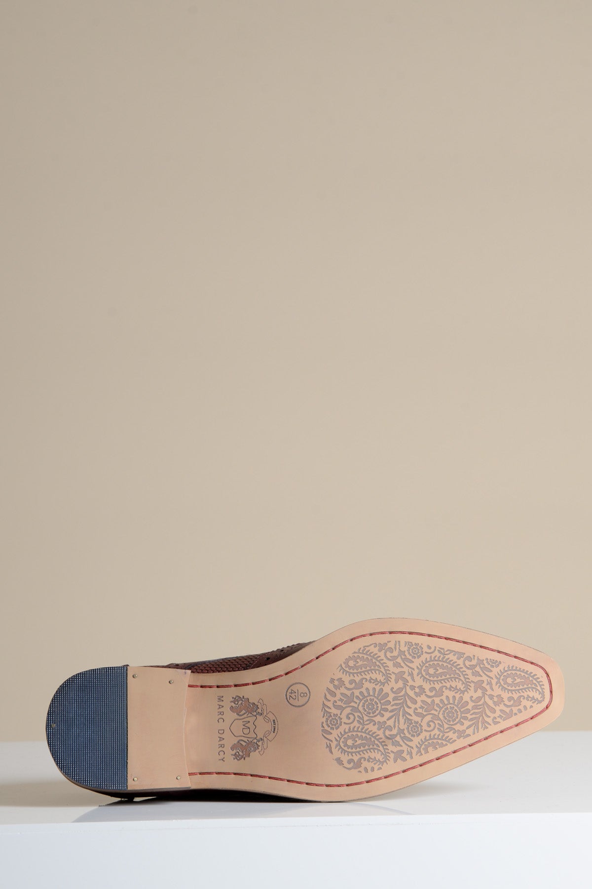 The marcdarcy BRANDON - Bordeaux Burgundy Leather Wingtip Brogue Shoe is shown on its side, highlighting the soles hand-crafted floral detailing and branded heel against a plain beige background.