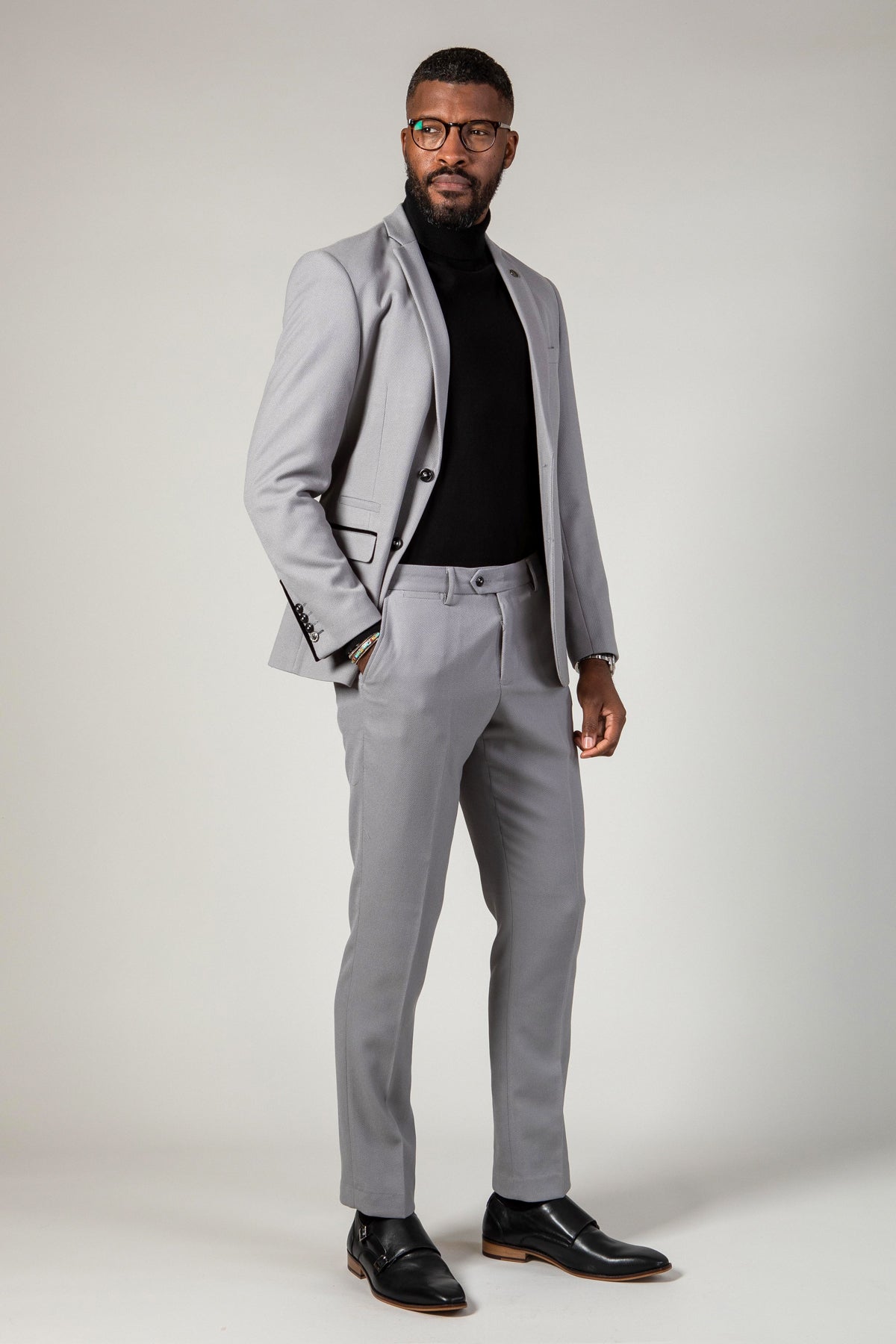 A Black man poses with his hand in his pocket, wearing the marcdarcy EDWIN Silver Grey Notch Lapel Two Piece Suit with a black turtleneck, black dress shoes, and glasses.