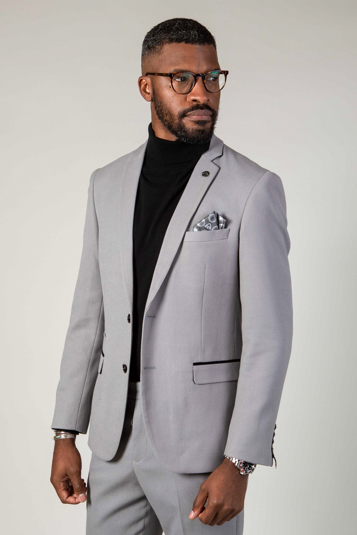 A Black man in the marcdarcy EDWIN Silver Grey Notch Lapel Two Piece Suit, paired with a black turtleneck and glasses, looks to the side against a plain background.