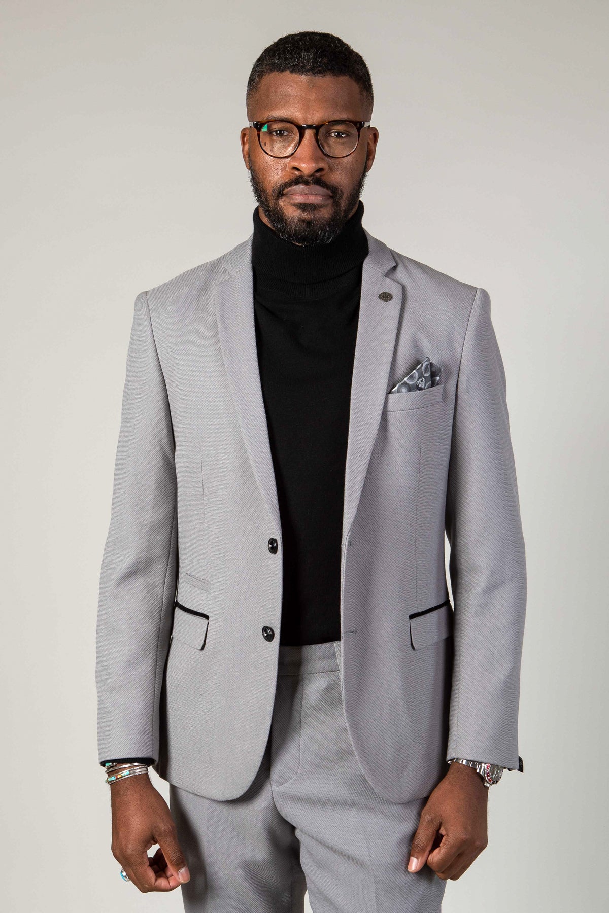 A man with short hair and a trimmed beard wears glasses and the marcdarcy EDWIN Silver Grey Notch Lapel Two Piece Suit with a pocket square and black turtleneck, standing against a plain light background.