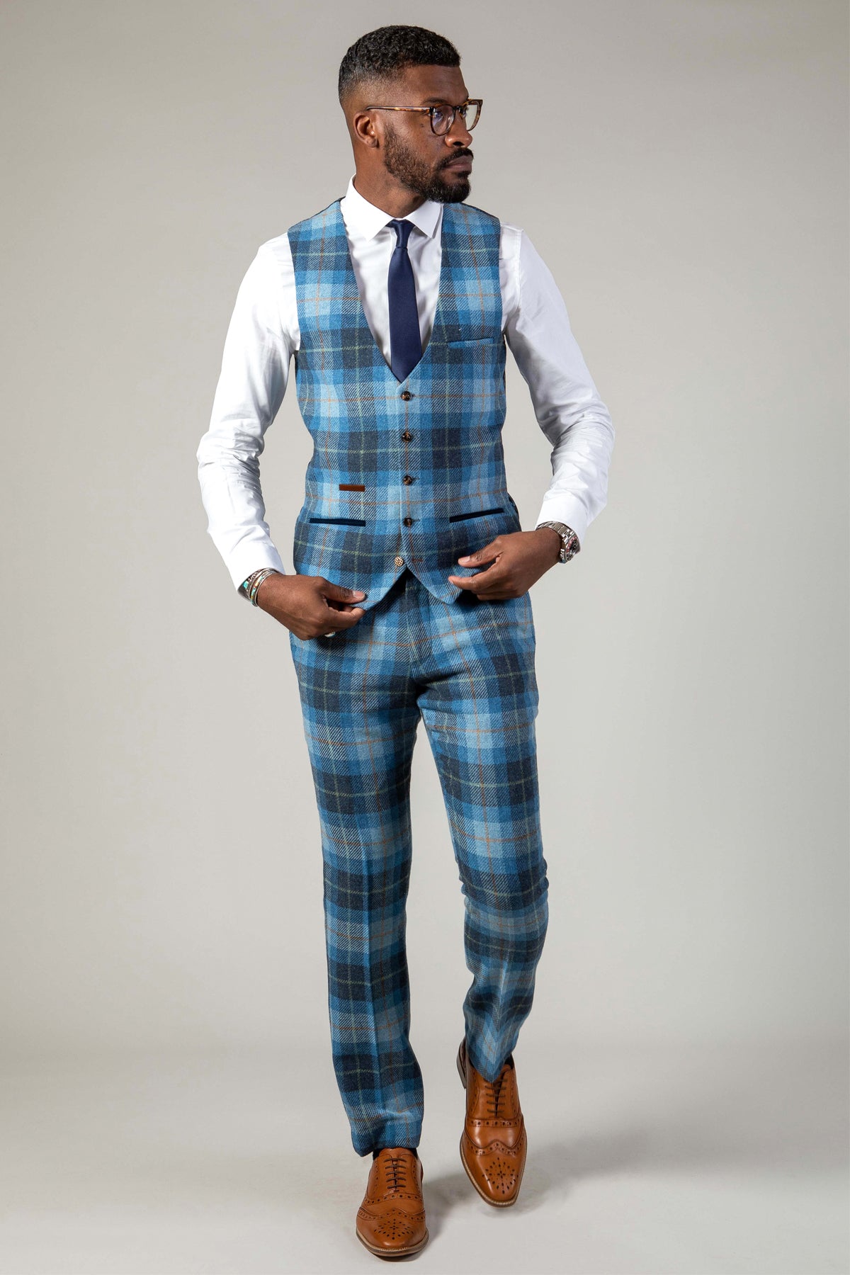 A man dressed in the Marc Darcy MORRIS Blue Tweed Check Waistcoat, styled with a white shirt, navy tie, and brown shoes, poses against a plain light background, gazing to the side with one hand in his pocket.