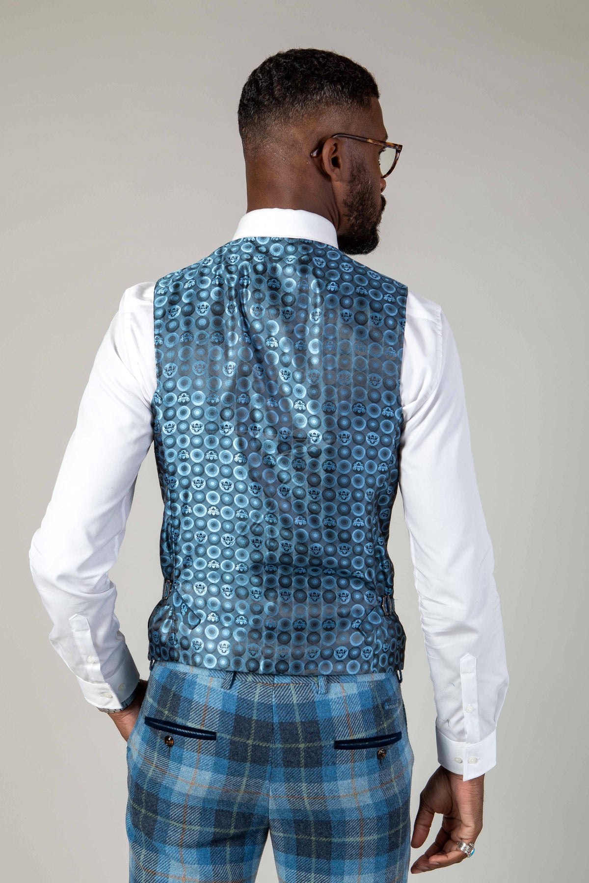 A man with short hair, beard, and glasses faces away, wearing a white shirt and the Marc Darcy MORRIS Blue Tweed Check Waistcoat with blue plaid trousers. The background is plain light gray.