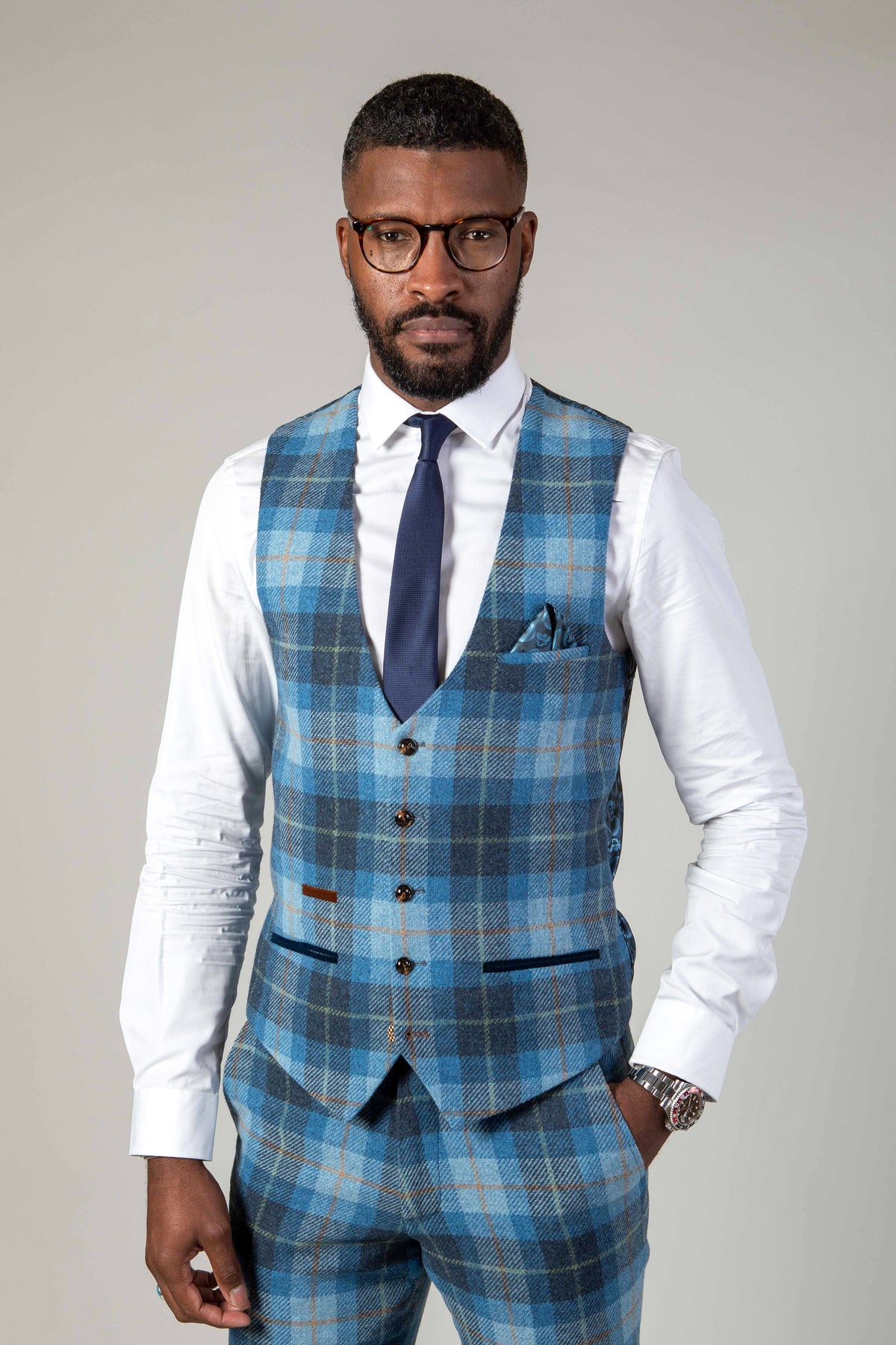 A man in glasses models the Marc Darcy MORRIS Blue Tweed Check Waistcoat with matching slim trousers, white dress shirt, navy tie, and pocket square, standing against a plain light background to showcase modern tailoring.