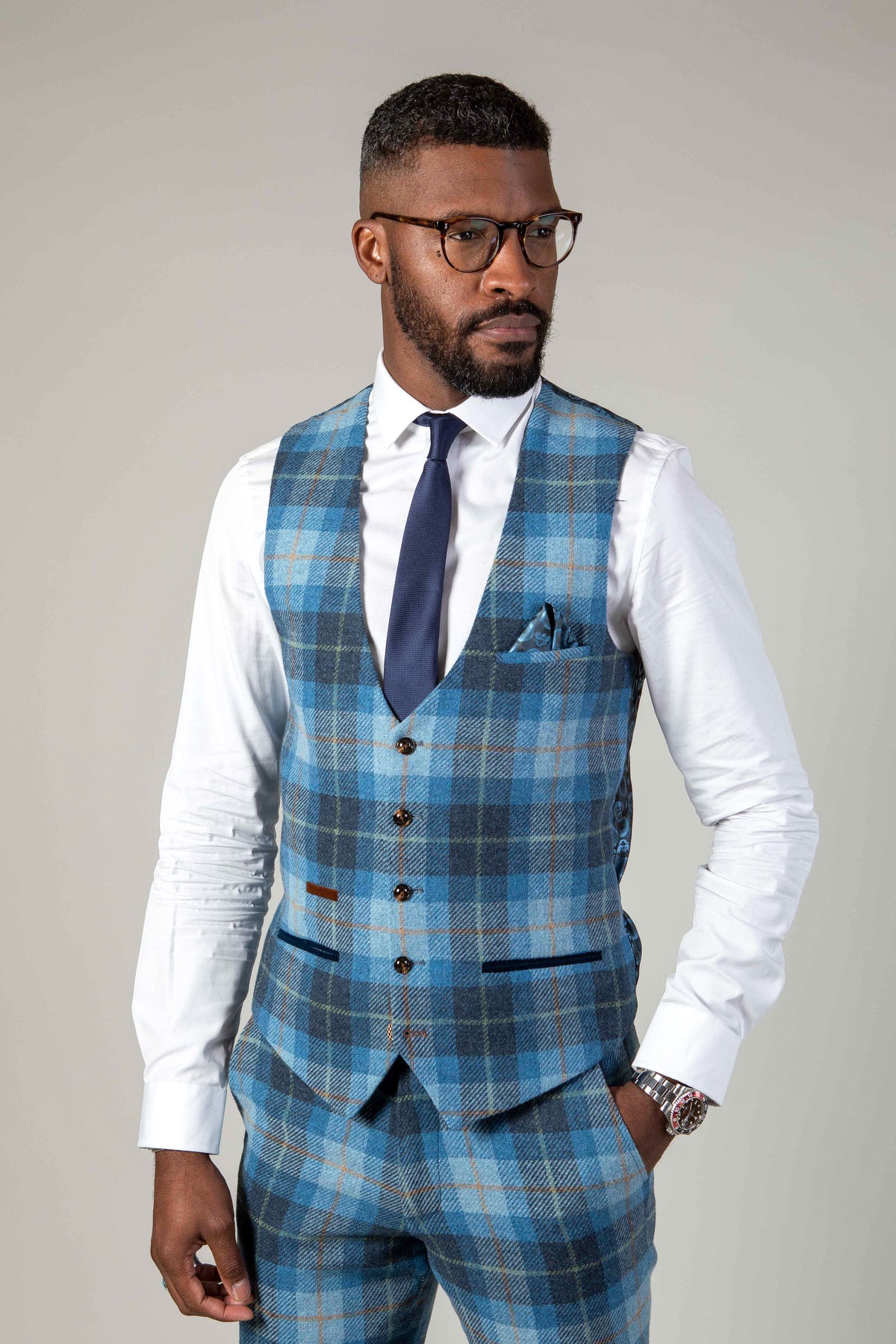 A Black man wears the Marc Darcy MORRIS Blue Tweed Check Waistcoat over a white shirt and navy tie, paired with glasses and a watch, showcasing modern tailoring in this slim fit look.