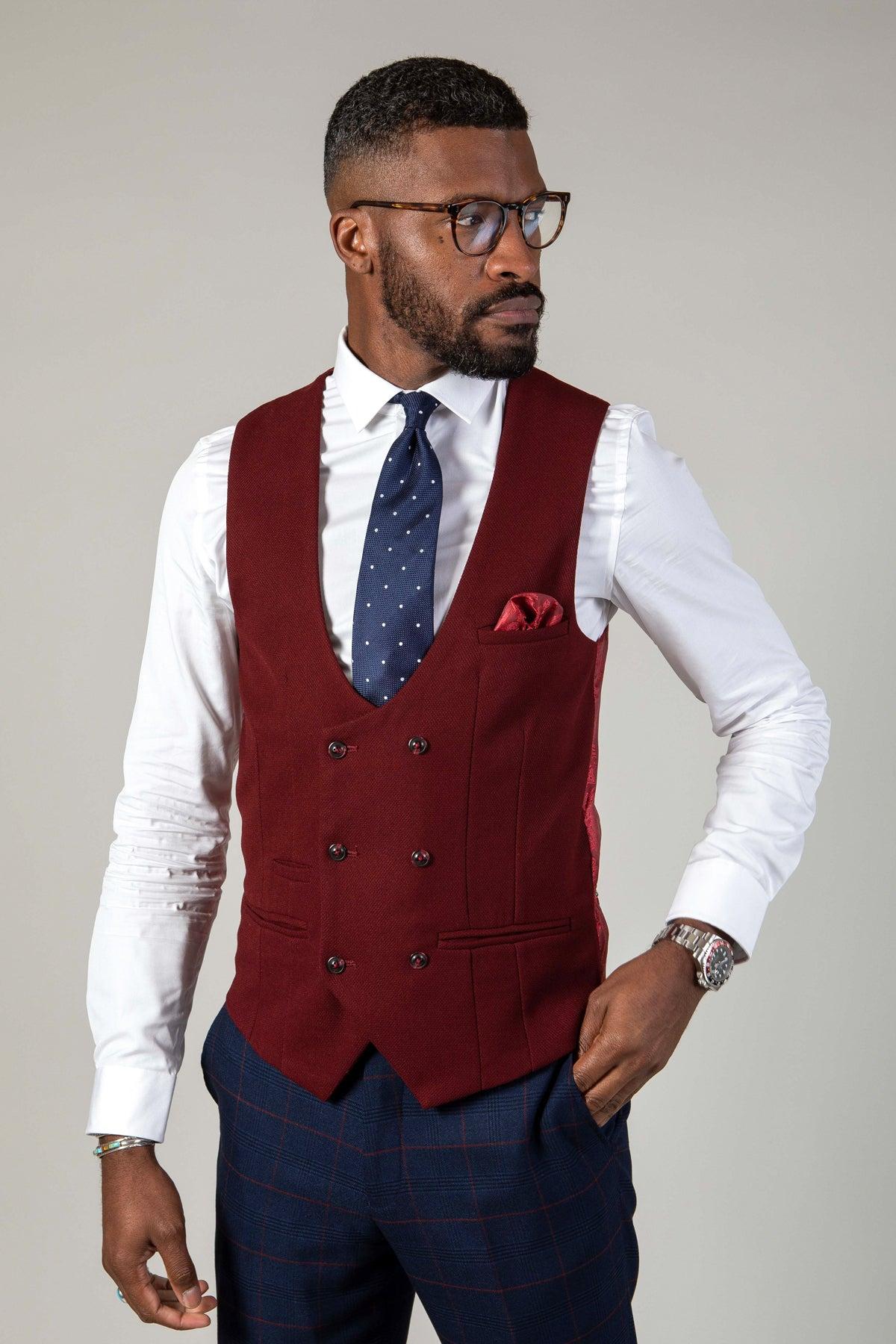 A man stands against a plain background wearing marcdarcy's KELVIN wine double-breasted waistcoat, glasses, a white dress shirt, navy blue polka-dot tie, matching pocket square, and blue plaid trousers—showcasing modern men’s suit style.