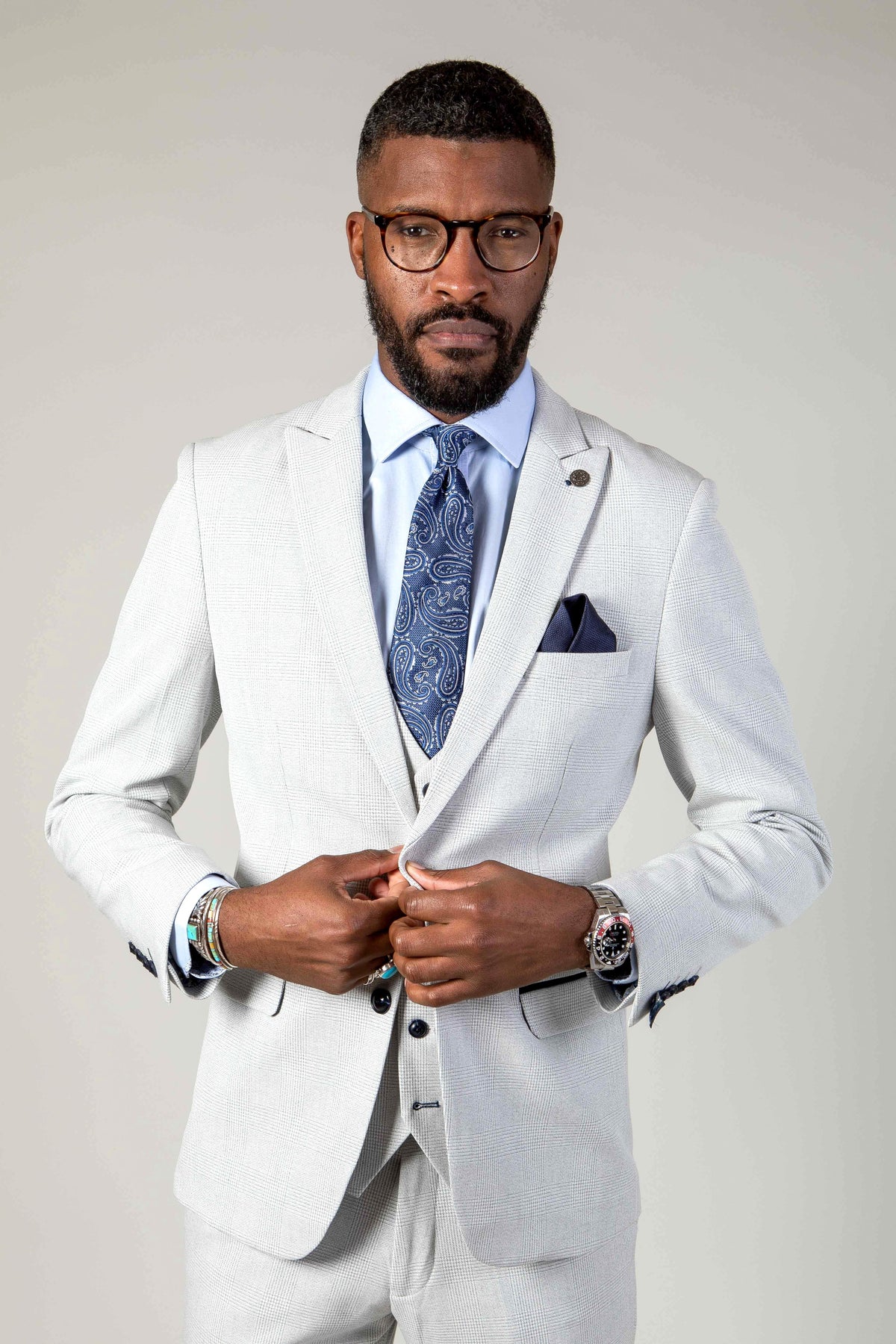 A man with a beard and glasses models the Marc Darcy BROMLEY Stone Check Suit with Kelvin Pink Waistcoat, wearing a light blue shirt and patterned blue tie. He buttons his jacket, accessorized with a watch and bracelets, against a plain light background.