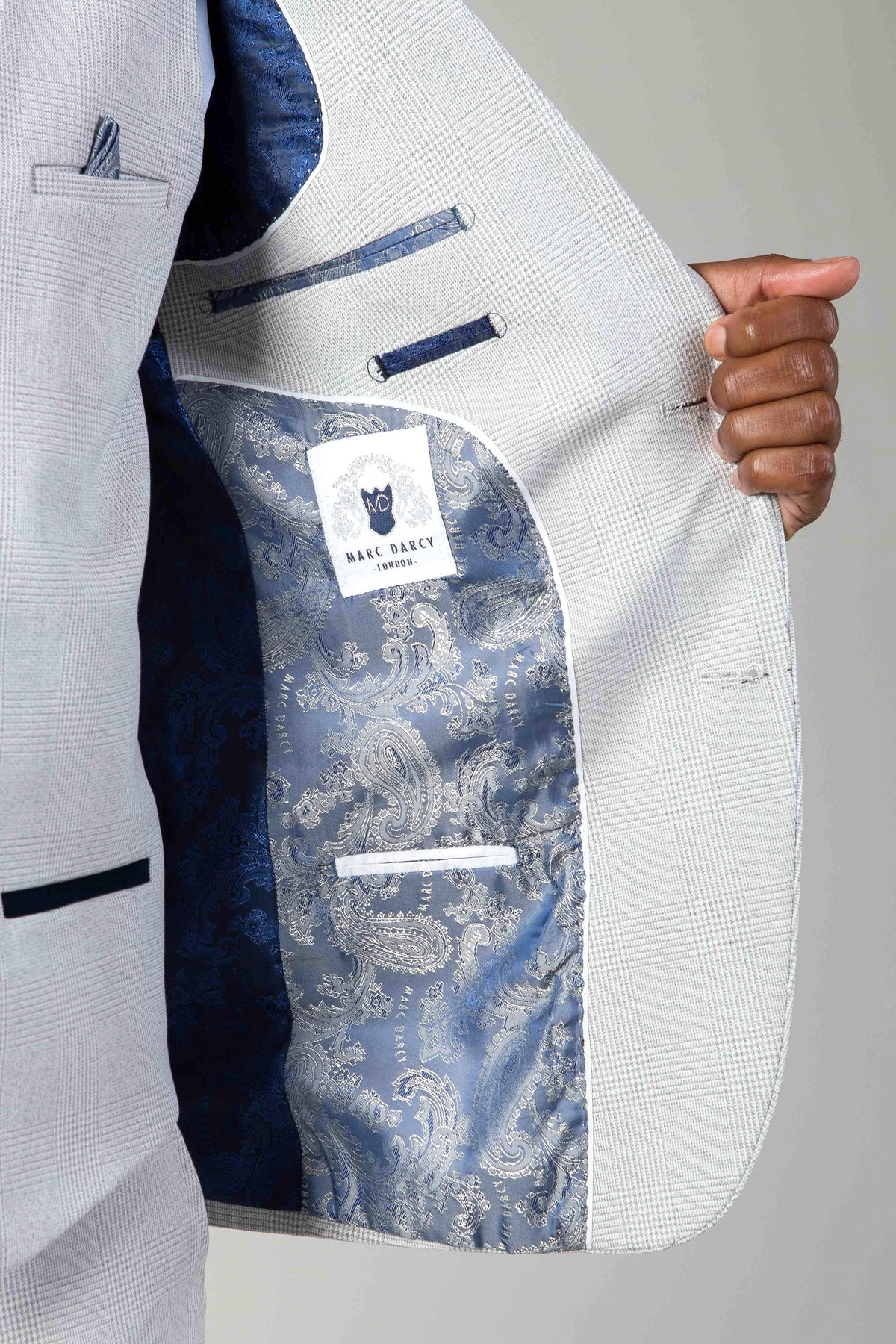 A person holds open a Marc Darcy BROMLEY Stone Check Suit jacket with a Kelvin Pink Waistcoat, revealing a blue paisley lining and a white label inside the inner pocket.