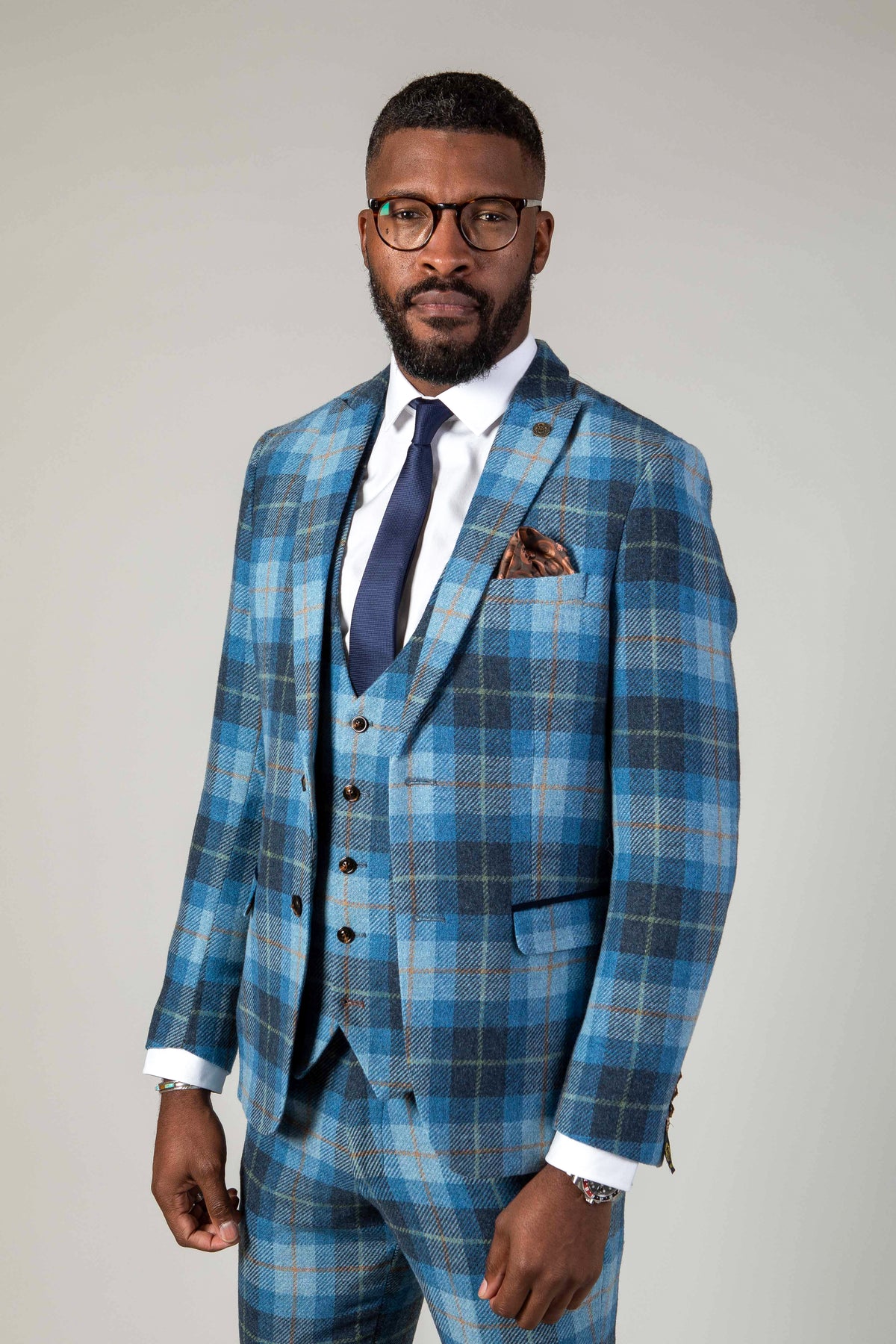 A Black man wearing the Marc Darcy MORRIS Blue Tweed Check Blazer, styled as a three-piece suit with a white shirt, navy tie, glasses, and pocket square, poses against a grey background.