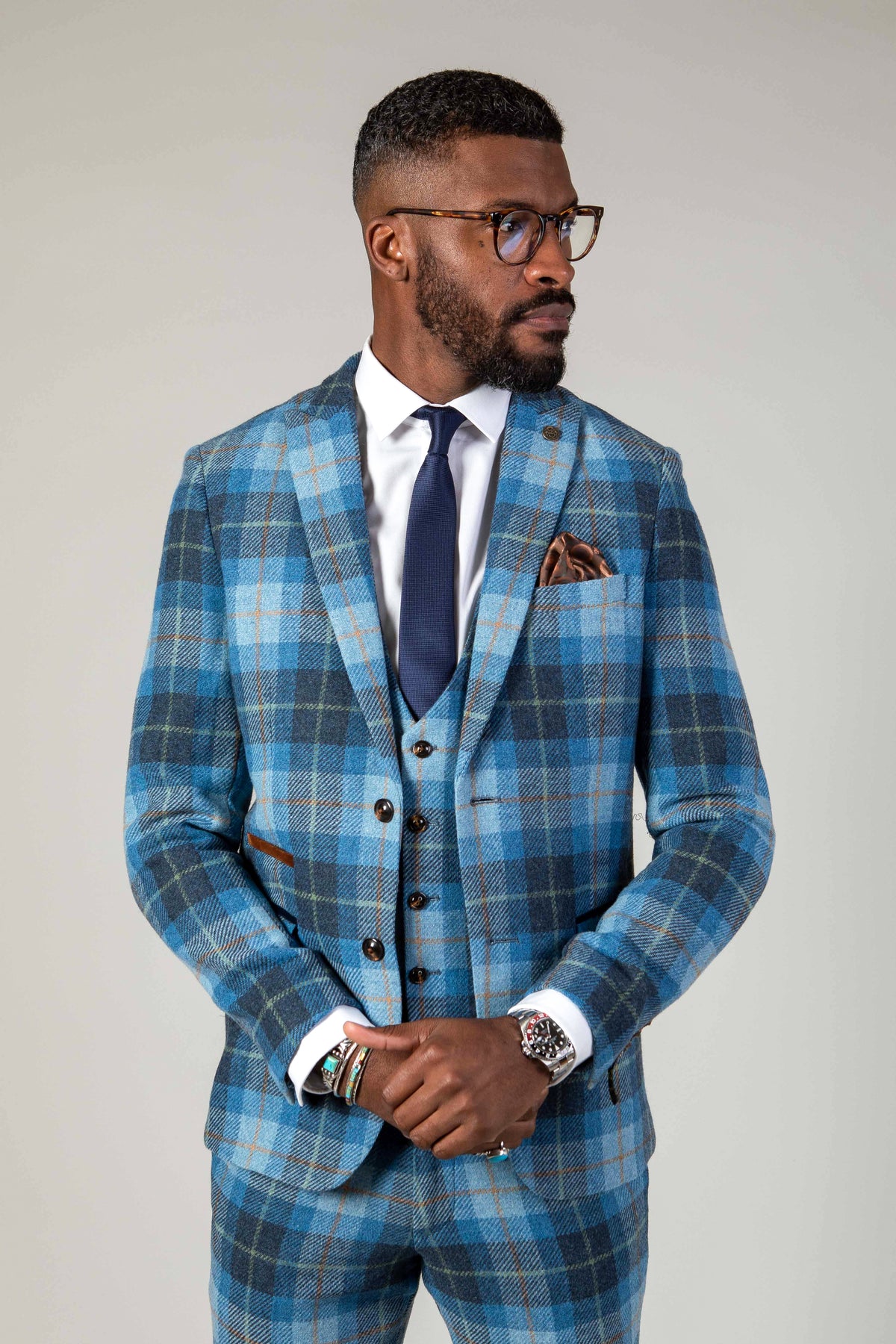 A man wearing glasses and the Marc Darcy MORRIS Blue Tweed Check Blazer with a white shirt and navy tie stands looking to his left, hands loosely clasped at his waist.
