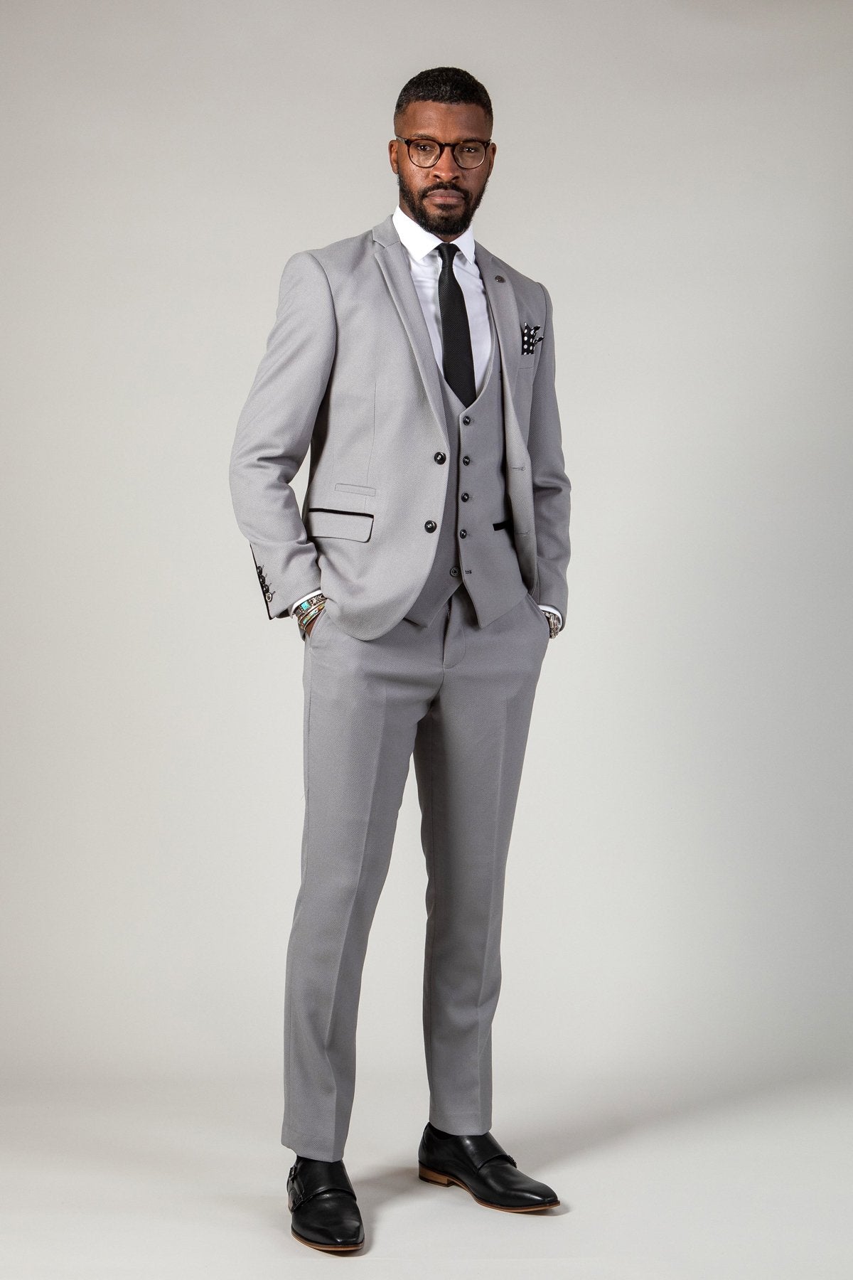 A bearded man with glasses stands confidently, hands in pockets, wearing the marcdarcy EDWIN Silver Grey Notch Lapel Three Piece Suit with a white shirt, black tie, and black dress shoes against a plain background.