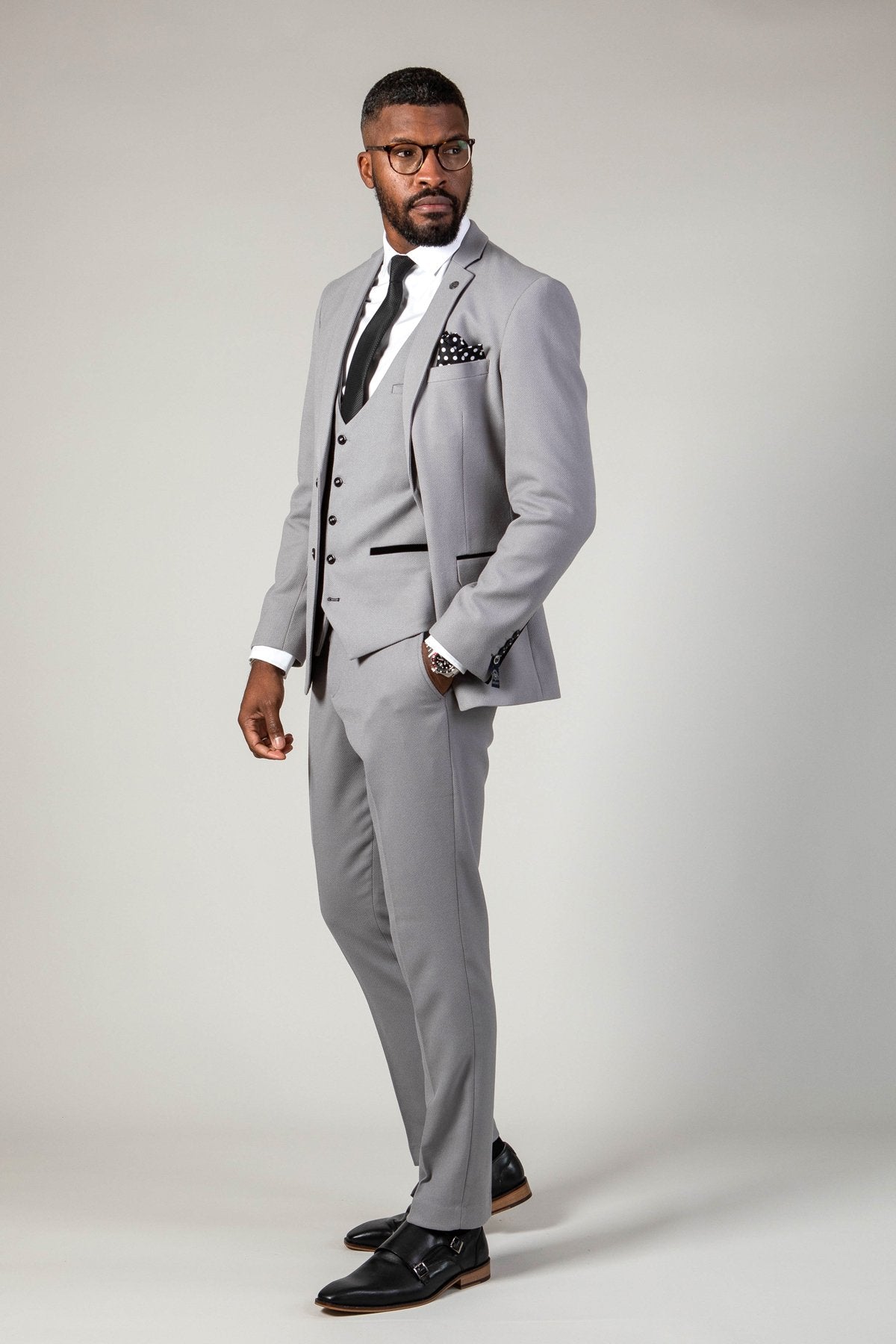 A man in the marcdarcy EDWIN Silver Grey Notch Lapel Three Piece Suit, with a white shirt, black tie, glasses, and a patterned pocket square stands with one hand in his pocket.