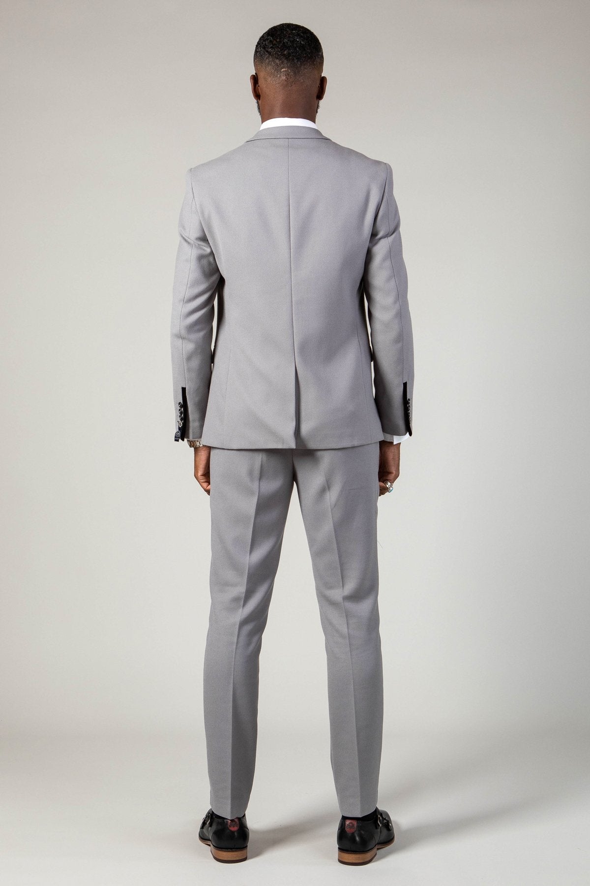 A person stands facing away from the camera, dressed in the marcdarcy EDWIN - Silver Grey Notch Lapel Three Piece Suit with black shoes, set against a plain light background.