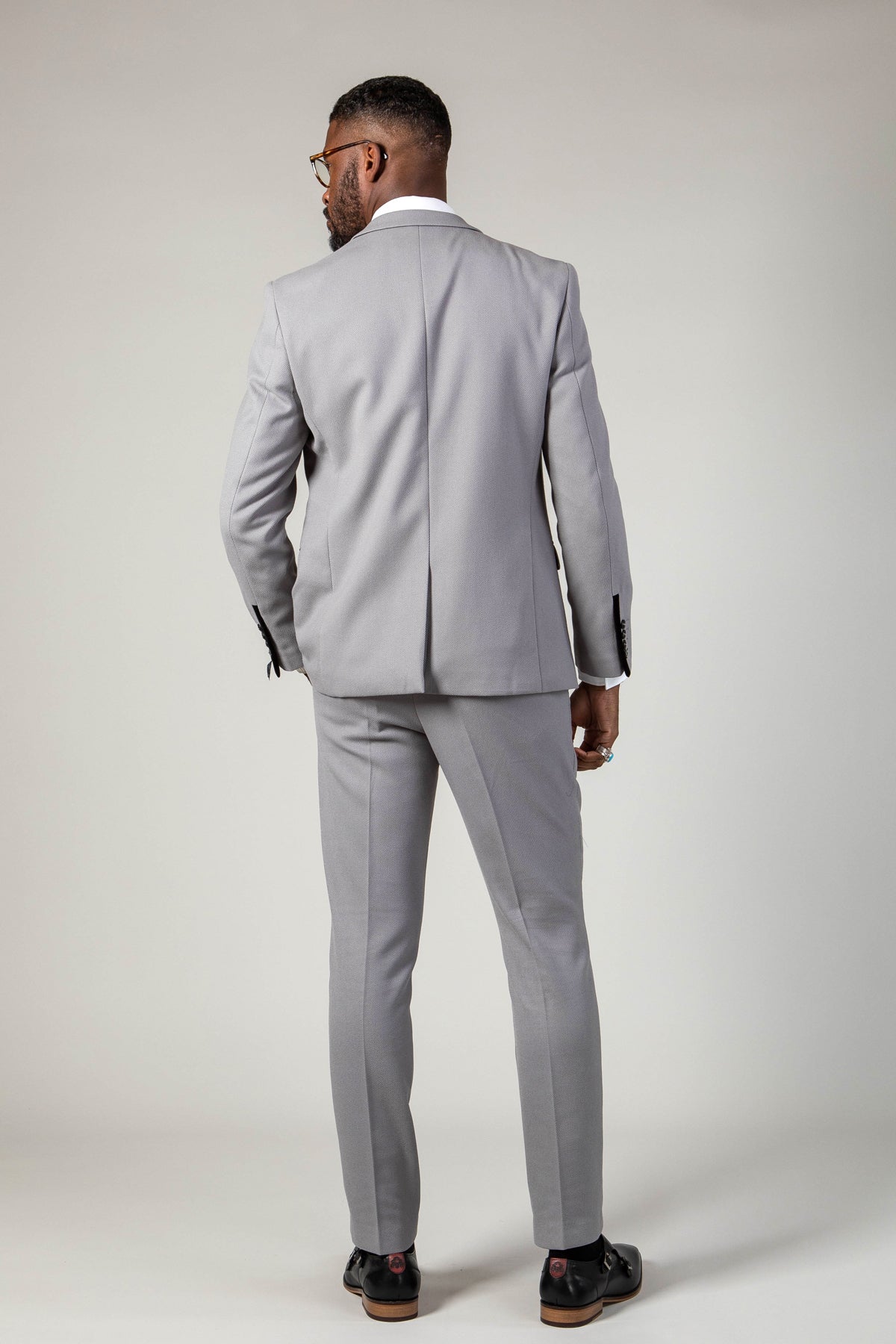 A man in the marcdarcy EDWIN Silver Grey Notch Lapel Two Piece Suit, paired with a white shirt and glasses, stands with his back to the camera wearing black shoes.