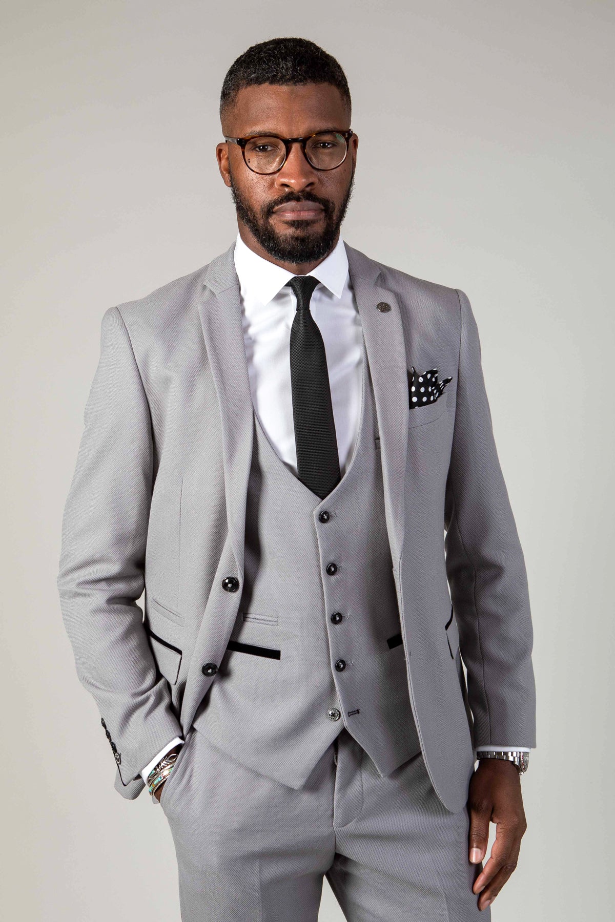 A man in the marcdarcy EDWIN - Silver Grey Blazer stands with his hand in his pocket, styled with a three-piece suit, white shirt, black tie, glasses, and a patterned pocket square.