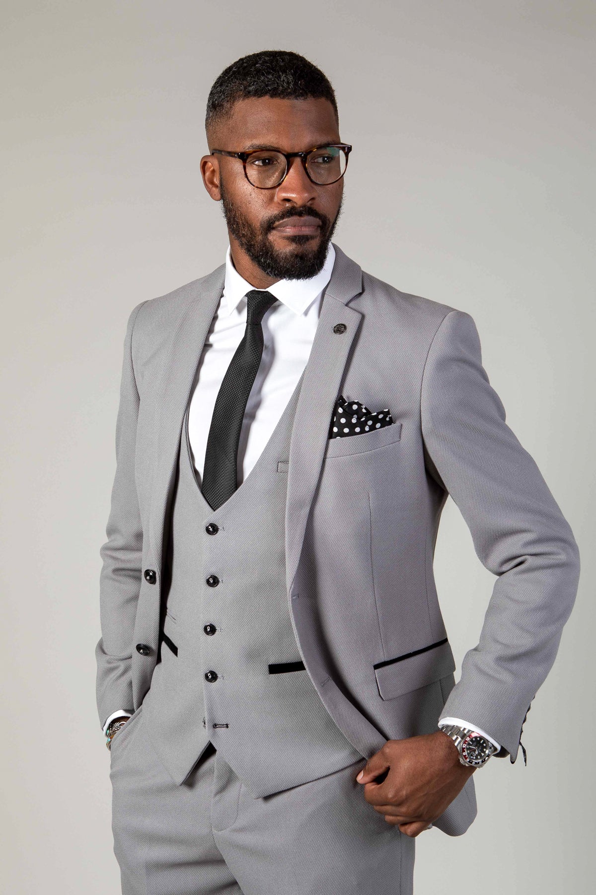 A Black man with a beard and glasses wears the marcdarcy EDWIN - Silver Grey Blazer in a slim fit, styled with a white shirt, black tie, polka dot pocket square, and a watch.