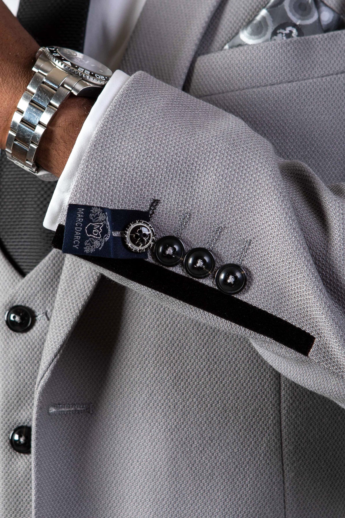 Close-up of EDWIN Silver Grey Notch Lapel Two Piece Suit by marcdarcy, highlighting the slim sleeve with black buttons, velvet trim, white shirt cuff, silver watch, and a navy marcdarcy label.