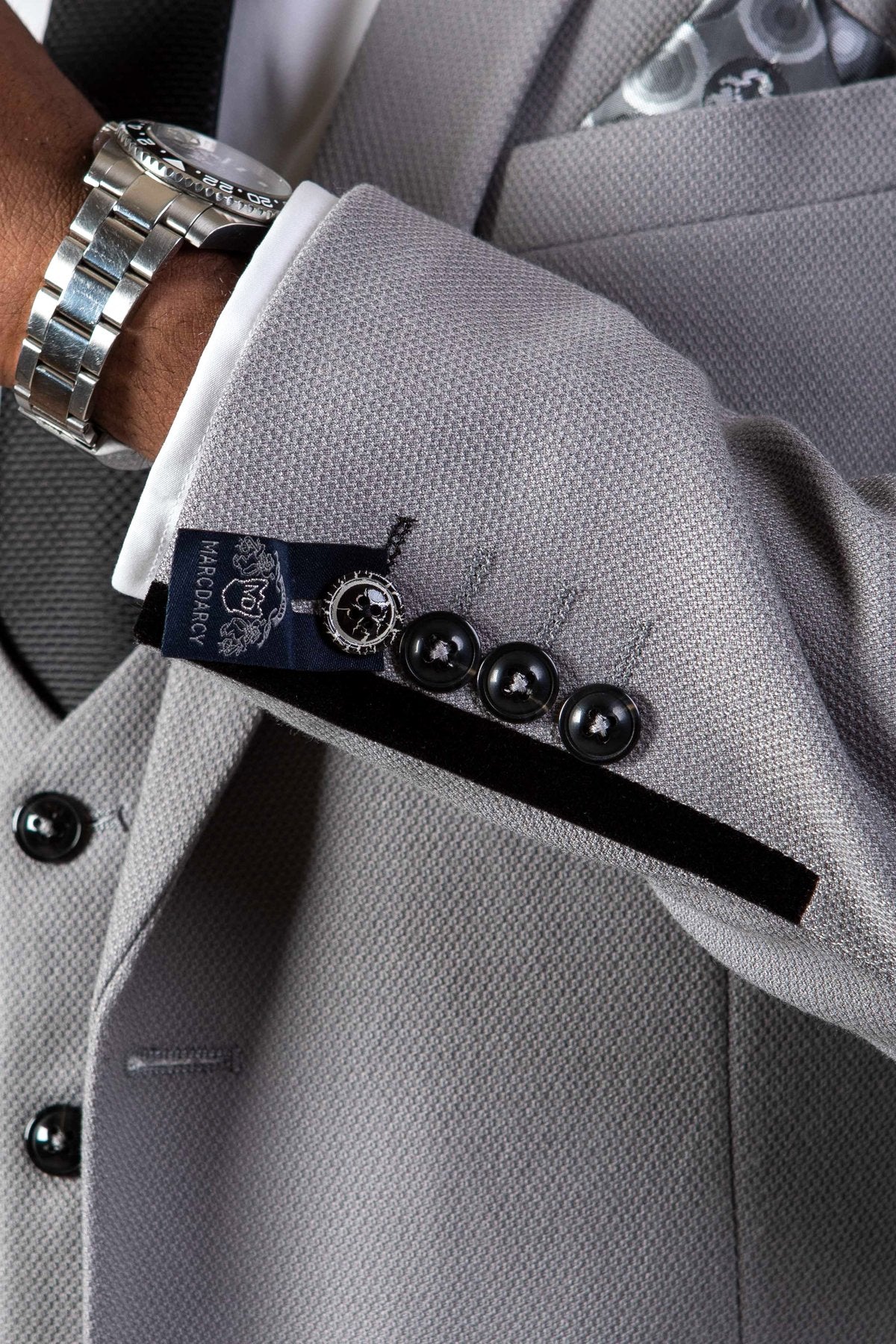 A close-up of a person wearing the marcdarcy EDWIN - Silver Grey Notch Lapel Three Piece Suit with a white shirt, patterned tie, silver watch, and slim fit blazer sleeve detailed with three black buttons and a designer label by the cuff.