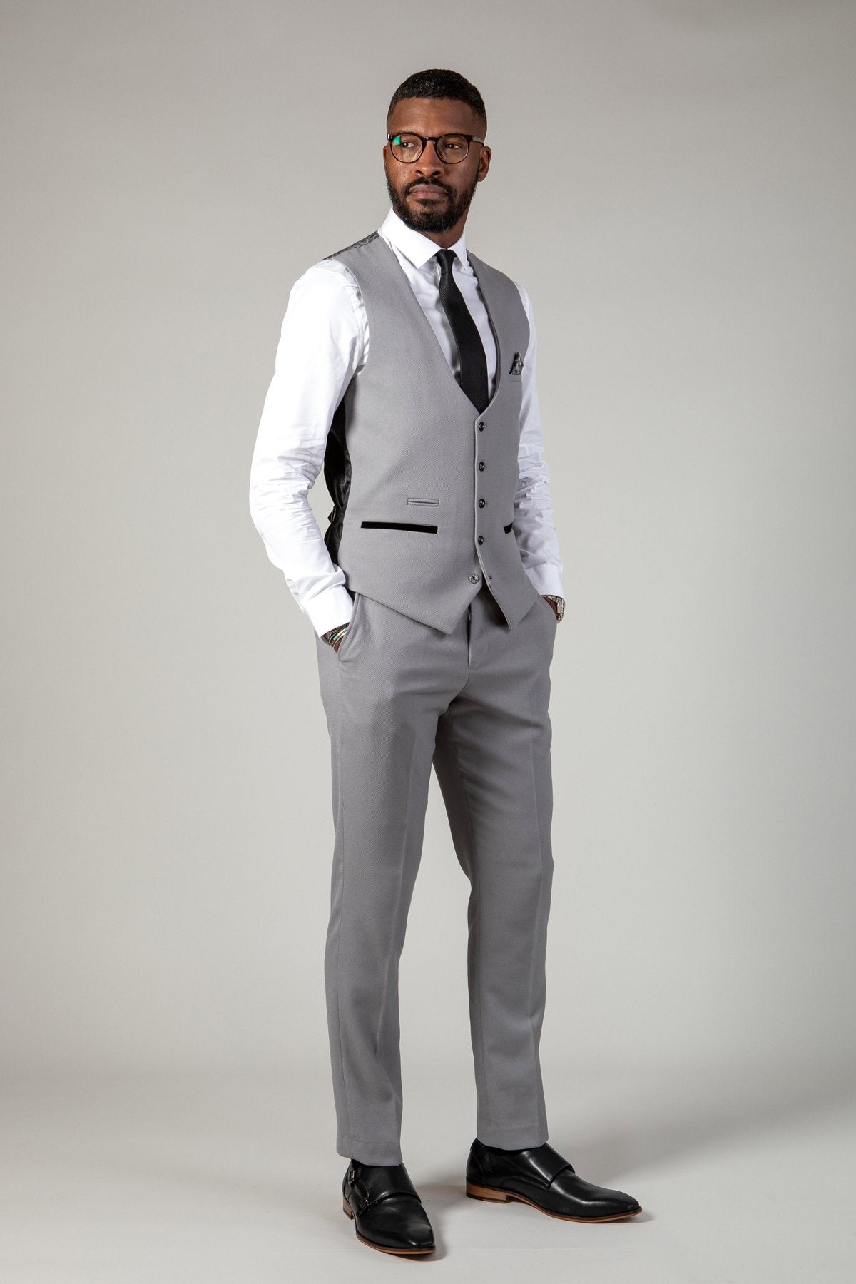 A man wears the marcdarcy EDWIN Silver Grey Notch Lapel Three Piece Suit with a slim fit blazer, white shirt, black tie, and black dress shoes. He stands hands in pockets, wearing glasses and looking to the side against a plain background.