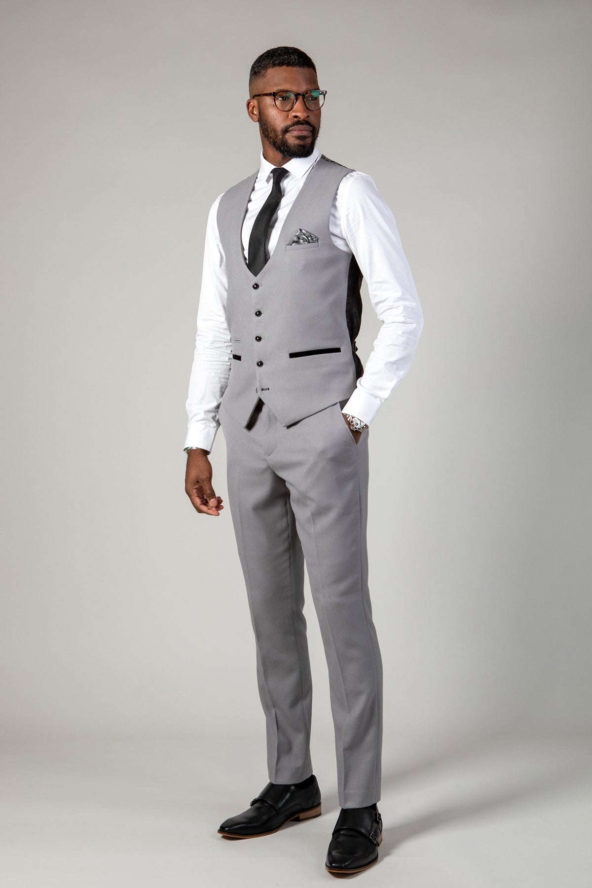 A man stands against a plain background wearing a light gray suit featuring the EDWIN - Silver Grey Single Breasted Waistcoat by Marc Darcy, along with a white shirt, black tie, pocket square, glasses, and black shoes, looking slightly to his left.