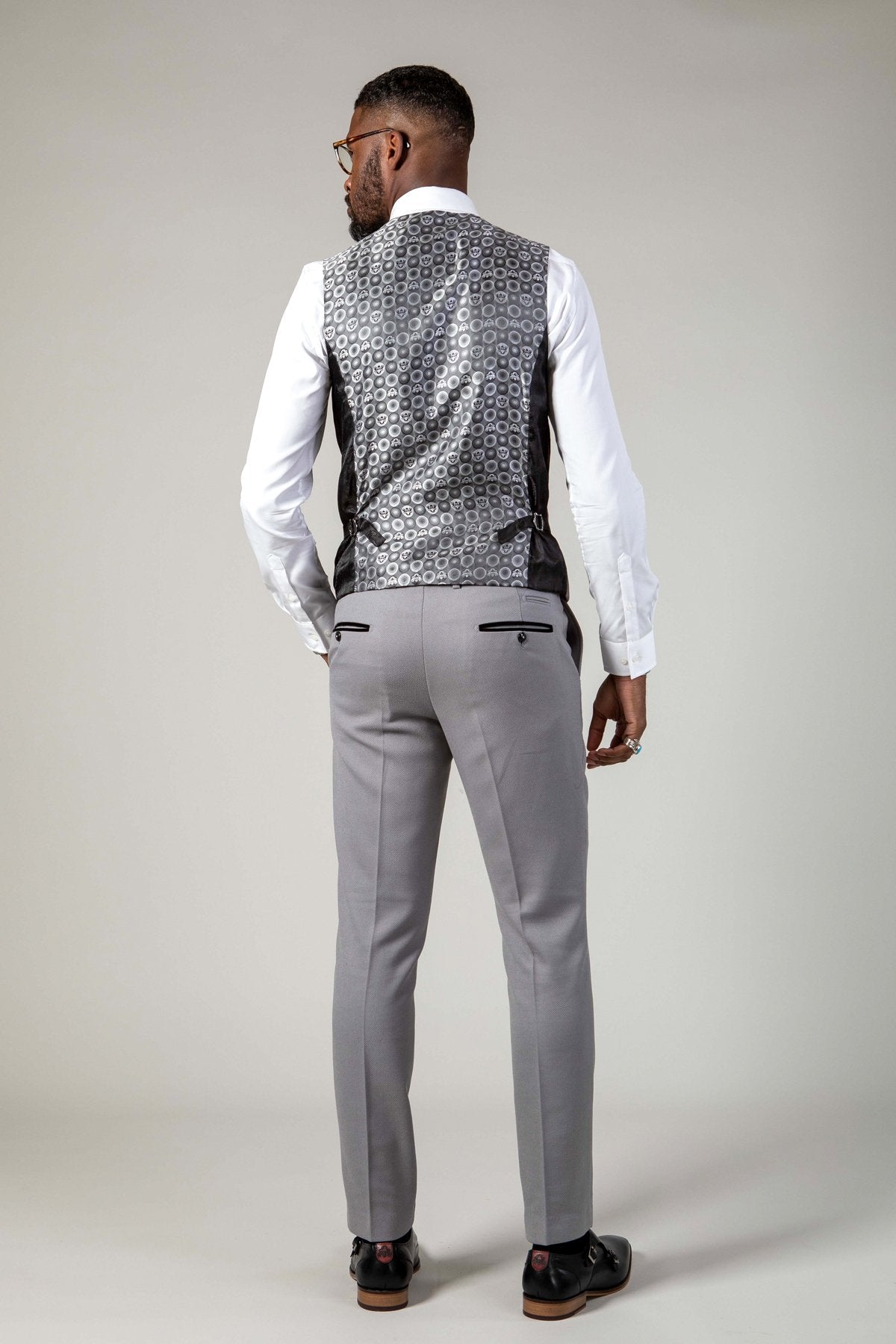 A man models the marcdarcy EDWIN - Silver Grey Notch Lapel Three Piece Suit, featuring a patterned vest and slim fit pants, paired with black shoes. He sports short hair, glasses, and a beard against a light background.