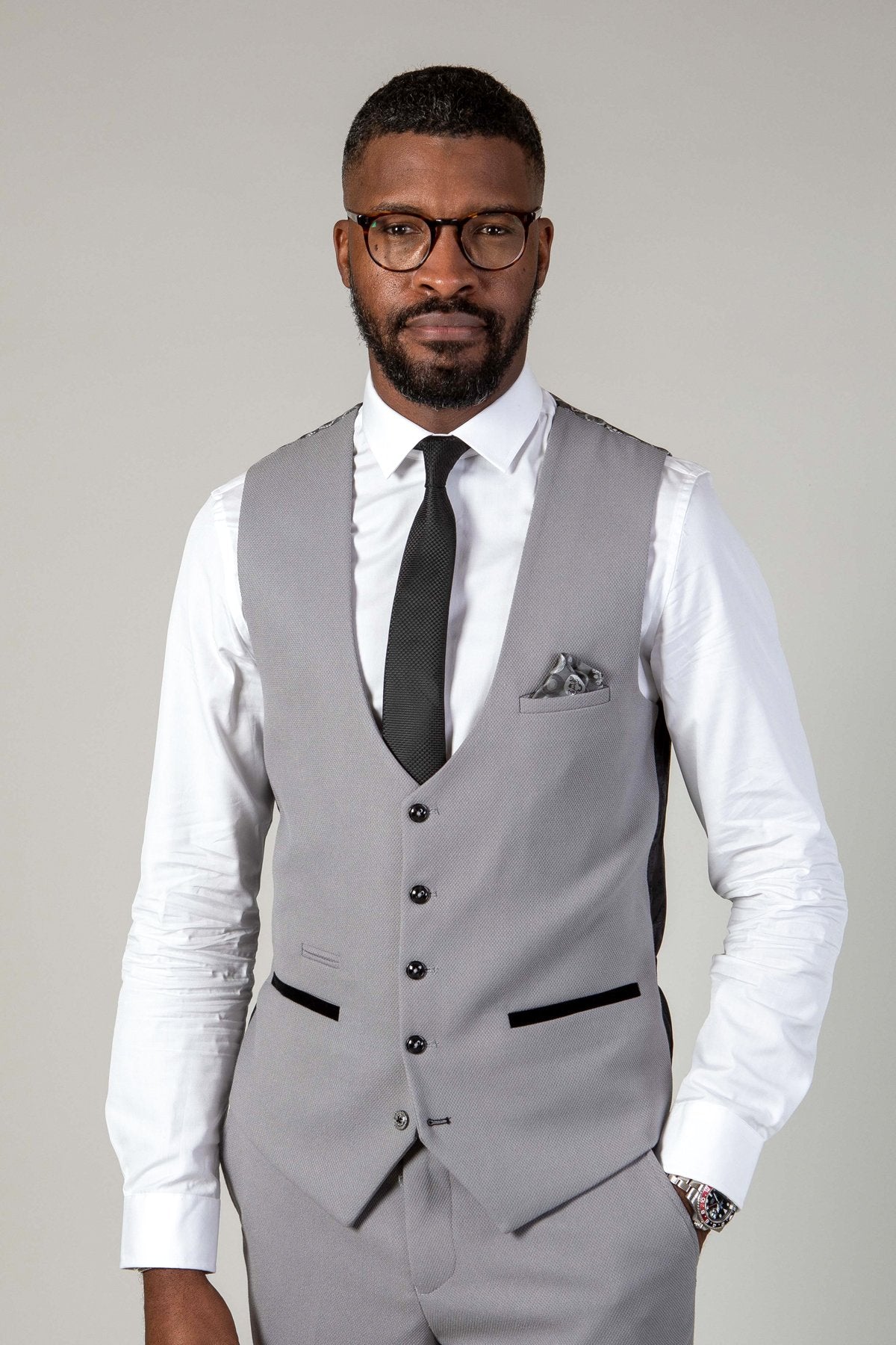 A Black man wearing glasses, a white dress shirt, black tie, and the marcdarcy EDWIN Silver Grey Notch Lapel Three Piece Suit stands with his hand in his pocket.