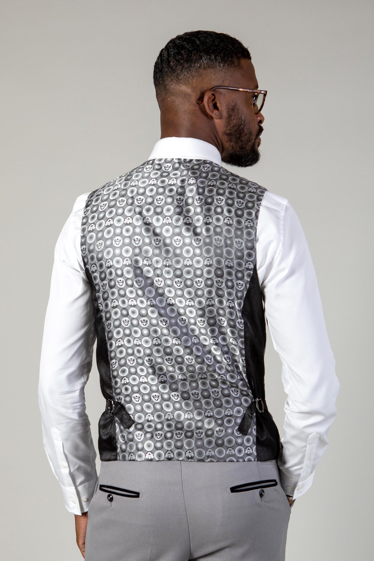 A man wearing a white dress shirt, gray pants, and the Marc Darcy EDWIN - Silver Grey Single Breasted Waistcoat featuring a slim fit with a repeating pattern of concentric circles and skull designs.