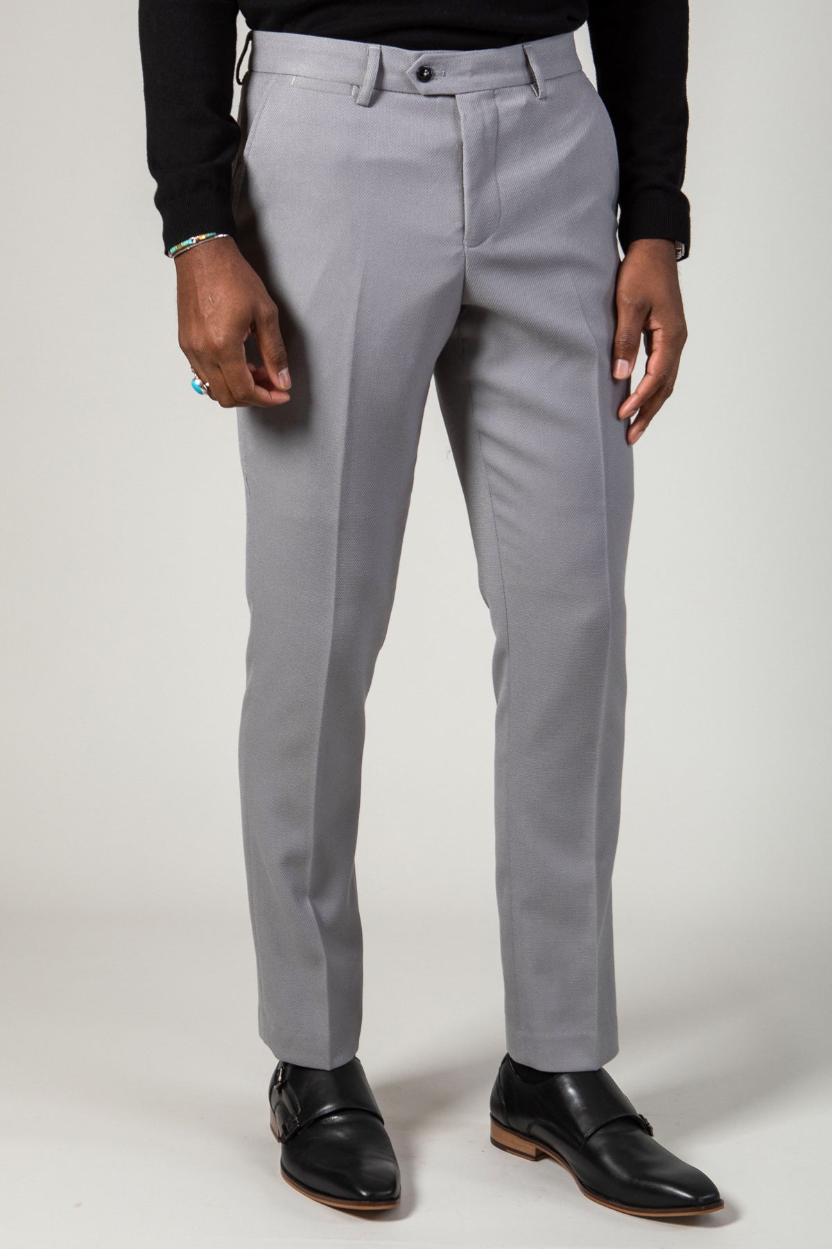 Close-up of a person wearing marcdarcy EDWIN - Silver Grey Trousers, black monk strap shoes, and a black sweater, accessorized with a beaded bracelet and ring on their left hand.