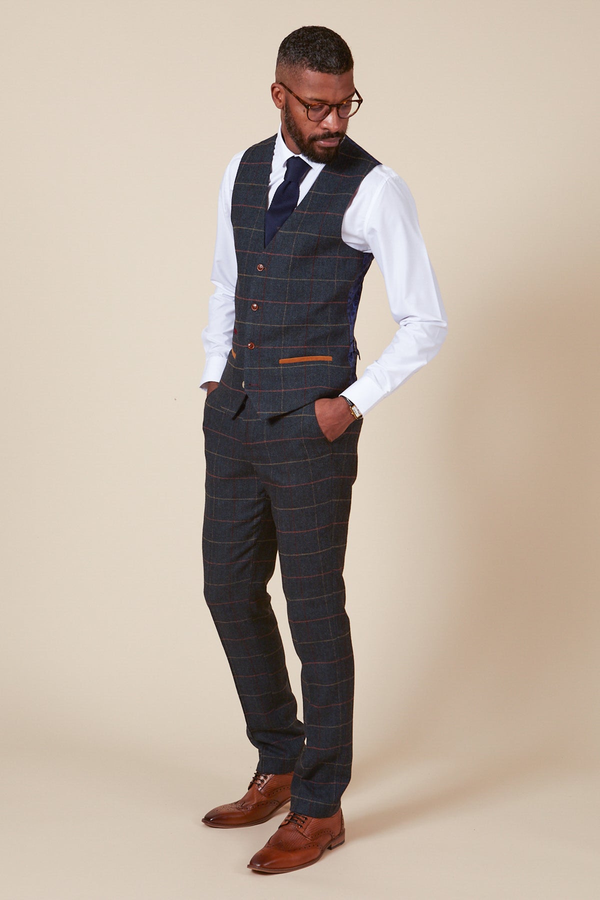 A man in a marcdarcy ETON single breasted navy blue tweed check waistcoat, dark plaid three-piece suit, white shirt, navy tie, glasses, and brown dress shoes stands with hands in pockets against a beige background.