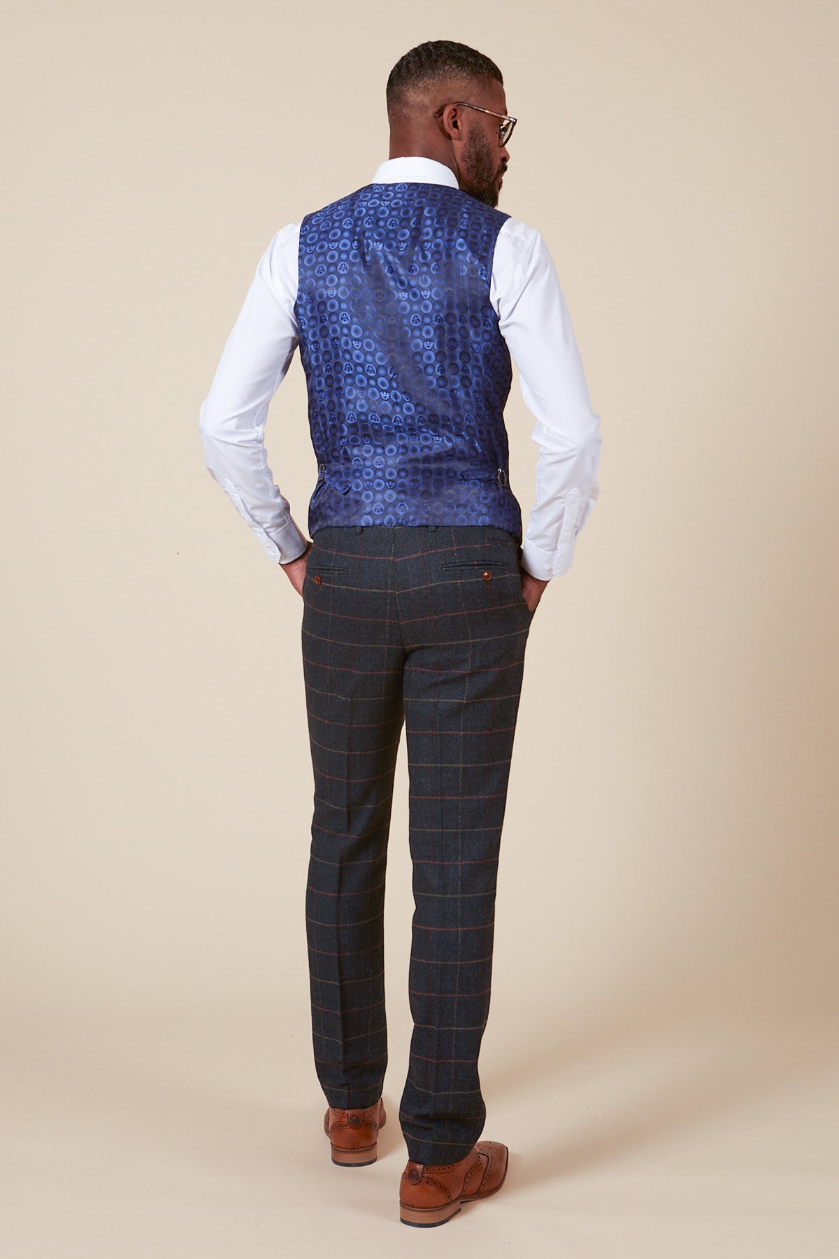A man in the marcdarcy ETON Single Breasted Navy Blue Tweed Check Waistcoat, white shirt, dark plaid trousers, brown belt, and brown shoes stands with hands in pockets and glasses against a plain beige background.