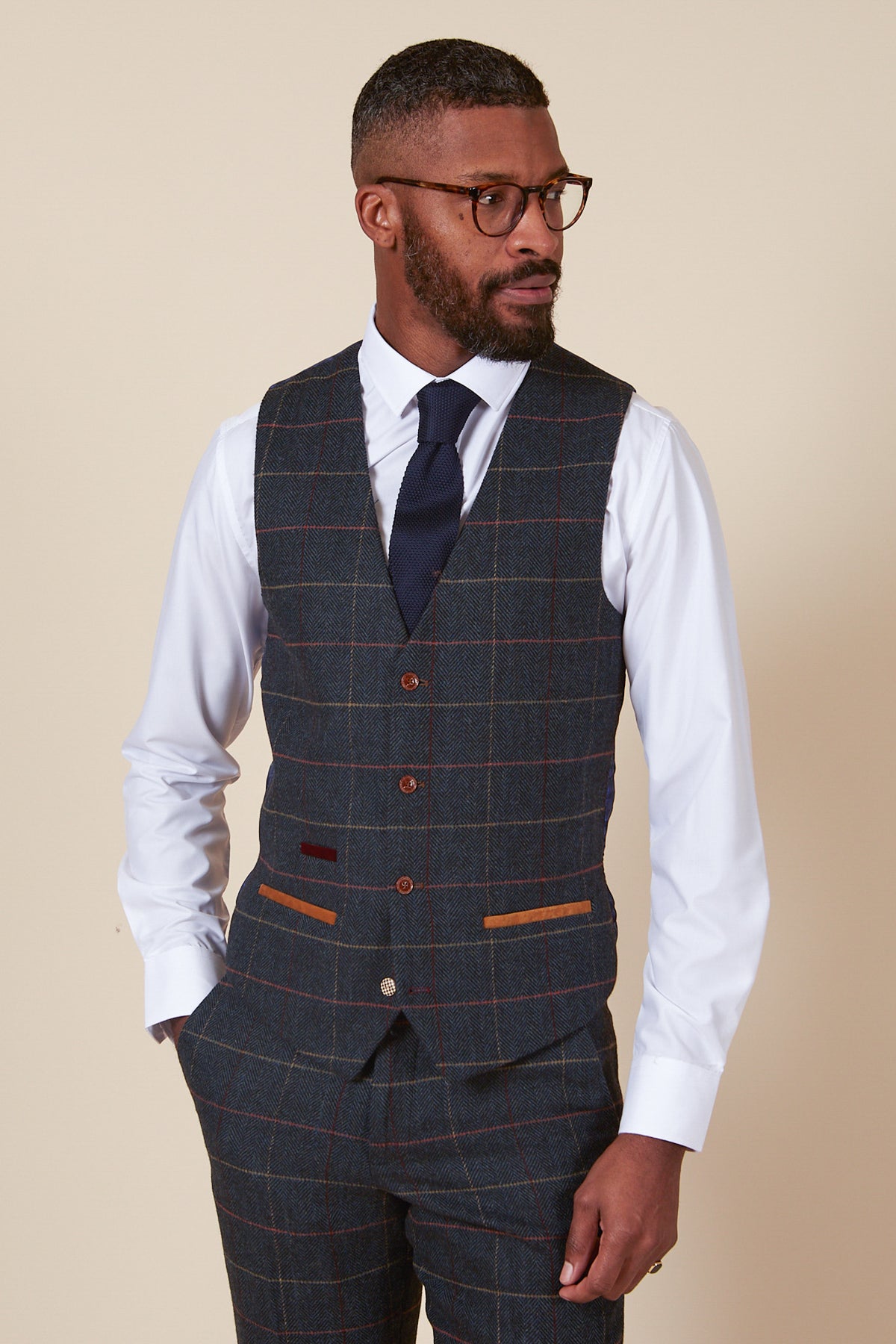 A Black man with a beard and glasses wears the marcdarcy ETON Single Breasted Navy Blue Tweed Check Waistcoat, paired with a white shirt and navy tie.