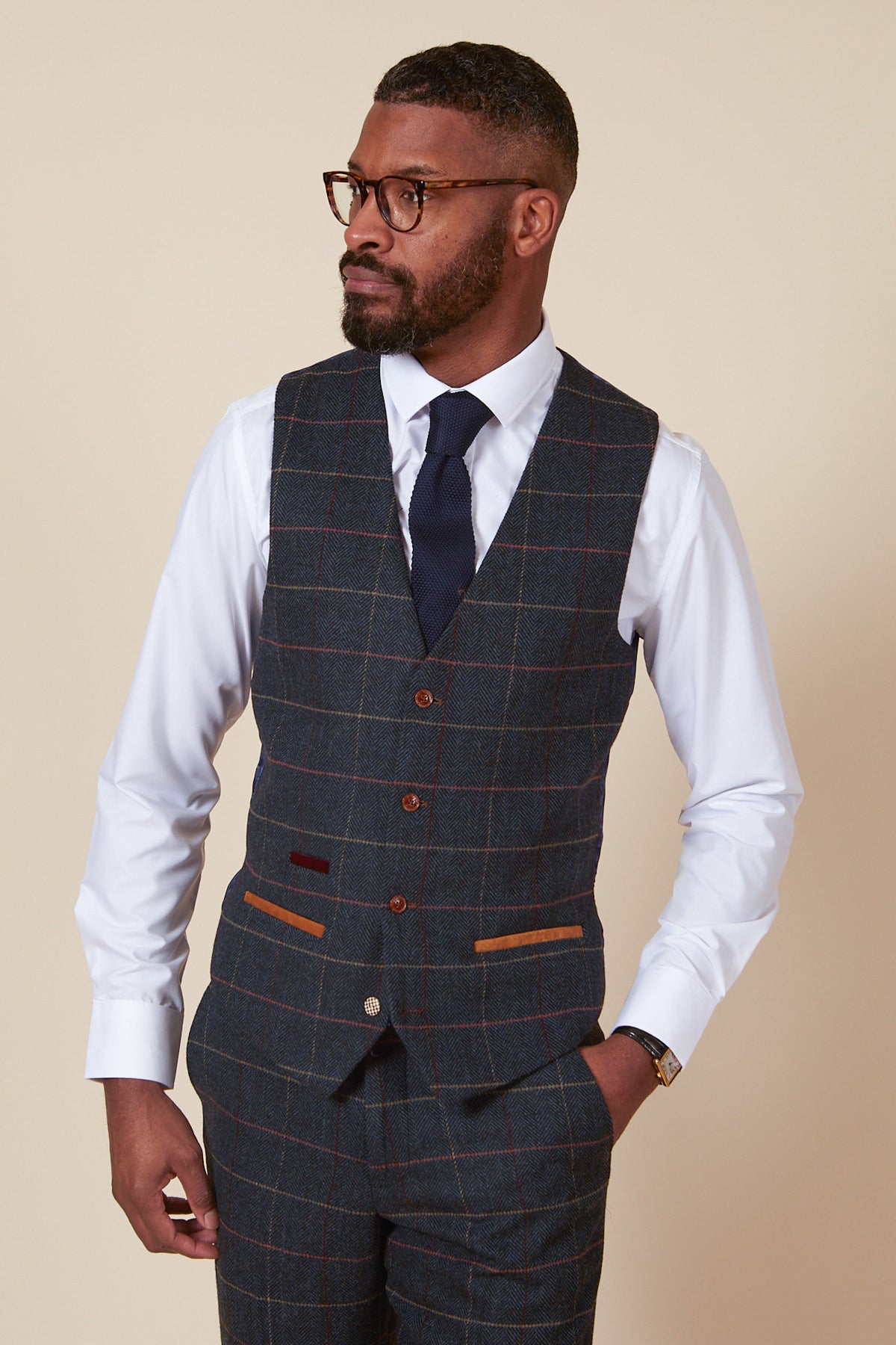 A man wears the marcdarcy ETON Single Breasted Navy Blue Tweed Check Waistcoat with tan pocket trims and brown buttons, styled with matching trousers, a white shirt, navy knit tie, and tortoiseshell glasses for a refined vintage look.