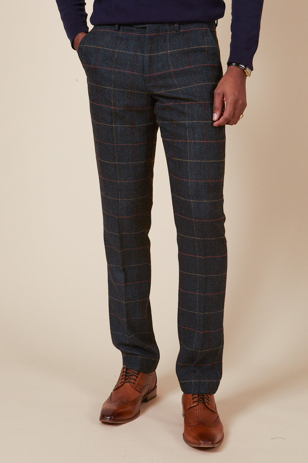 Against a beige backdrop, an individual stands in navy blue tweed check trousers by marcdarcy and brown leather dress shoes. Their hands rest in their pockets while wearing a dark long-sleeve top.