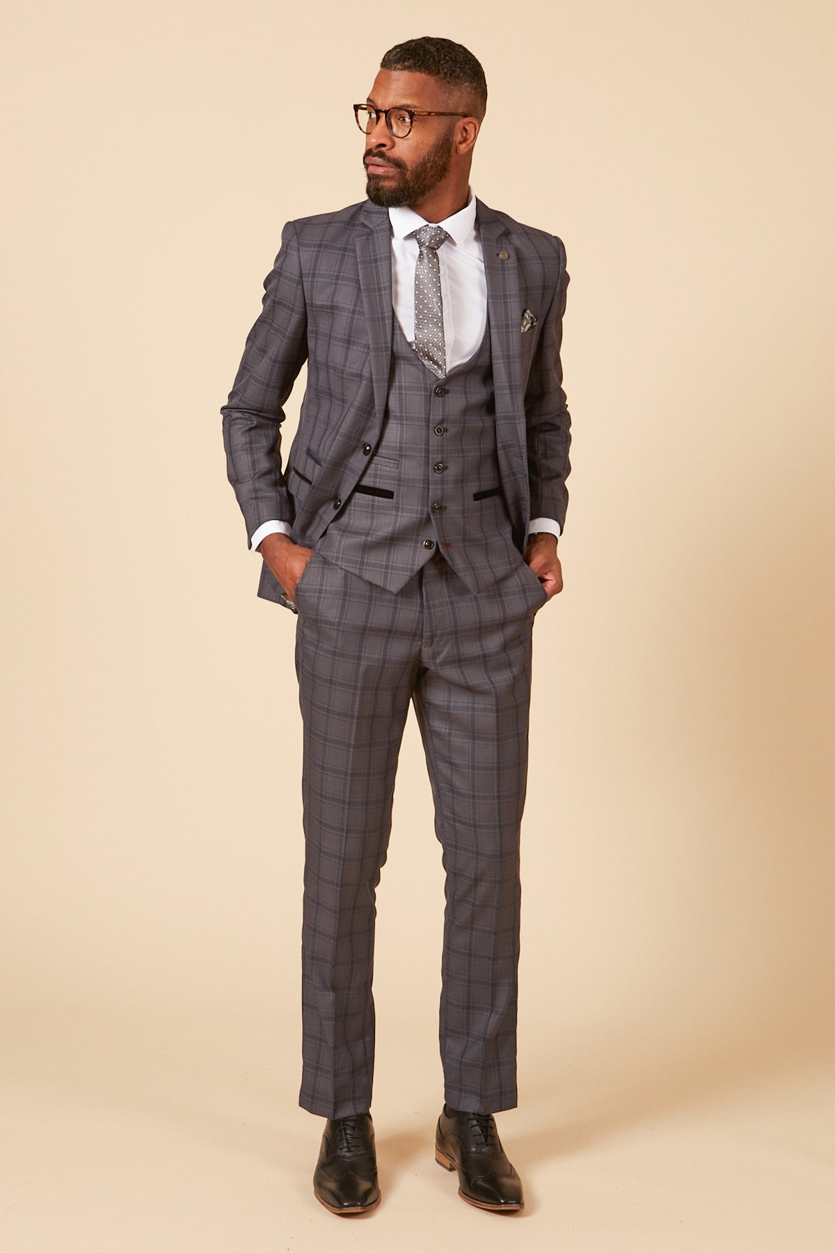 A man wears the marcdarcy JOSE grey check three piece suit with a white dress shirt and patterned tie, standing with hands in his pockets against a plain light background.