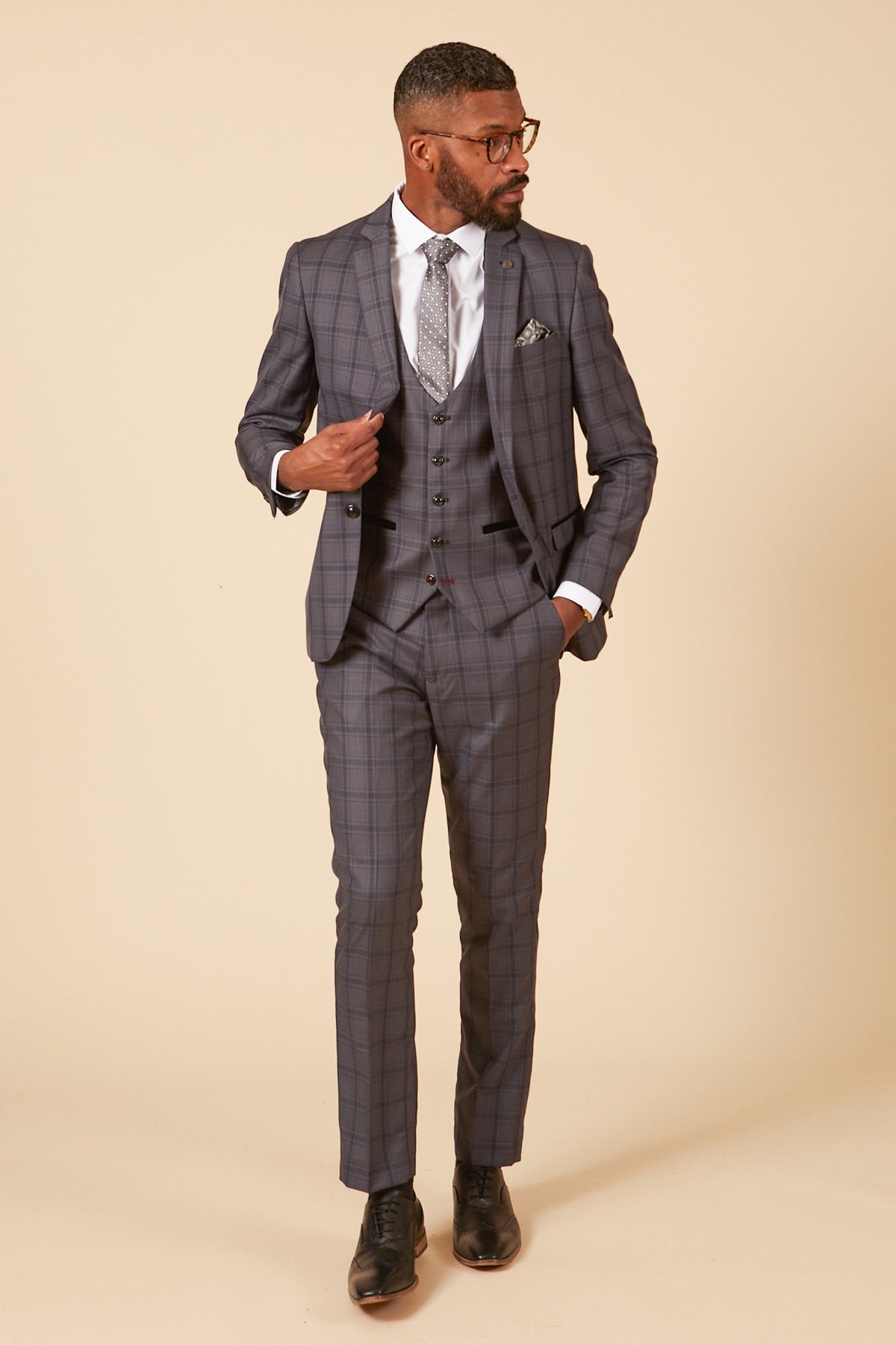 A man wearing the marcdarcy JOSE Grey Check Three Piece Suit with a white shirt, patterned tie, and black shoes stands against a beige background, looking to the side with one hand in his pocket and the other adjusting his jacket.