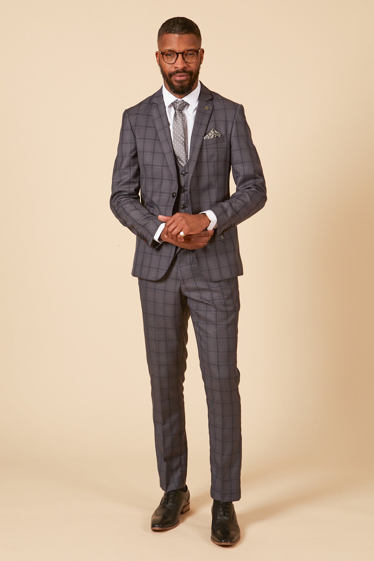 A man with short hair, beard, glasses, and pocket square poses against a beige backdrop in the marcdarcy JOSE - Grey Check Three Piece Suit with matching vest, white shirt, patterned tie, and black dress shoes.