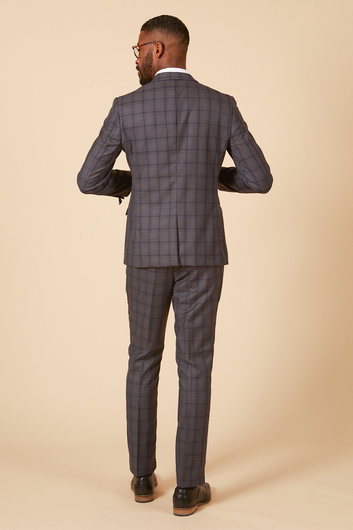 A man in the marcdarcy JOSE Grey Check Three Piece Suit, a tailored fit, with a white shirt and dark shoes stands facing away from the camera against a plain beige background.