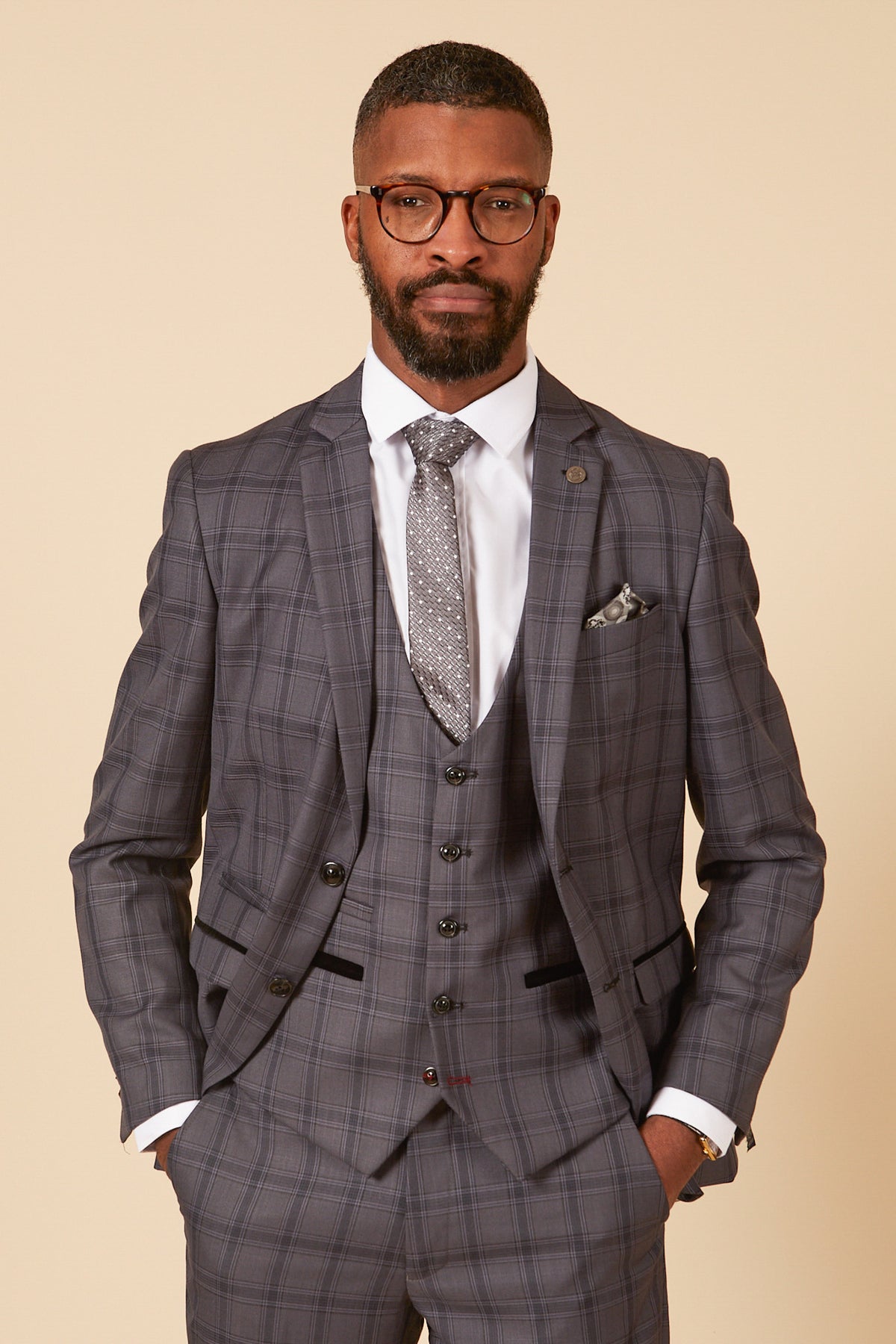 A man with short hair, a beard, and glasses wears the marcdarcy JOSE Grey Check Three Piece Suit in a tailored fit with a white shirt, patterned tie, and pocket square, standing with hands in pockets against a plain beige background.
