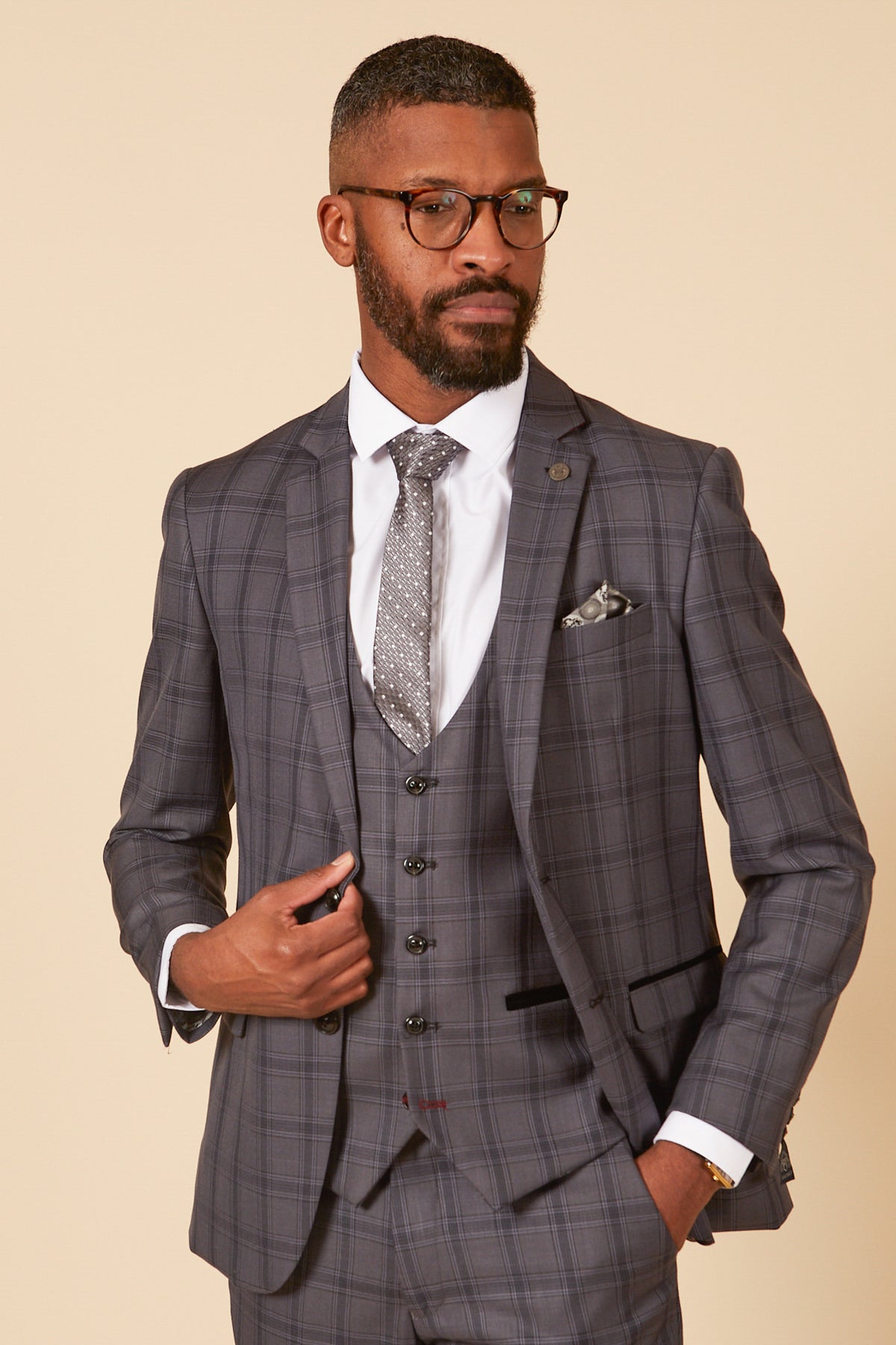 A man with short hair, a beard, and glasses wears the marcdarcy JOSE - Grey Check Three Piece Suit with a white shirt, patterned tie, and pocket square. He stands against a beige background, holding his jacket with one hand.
