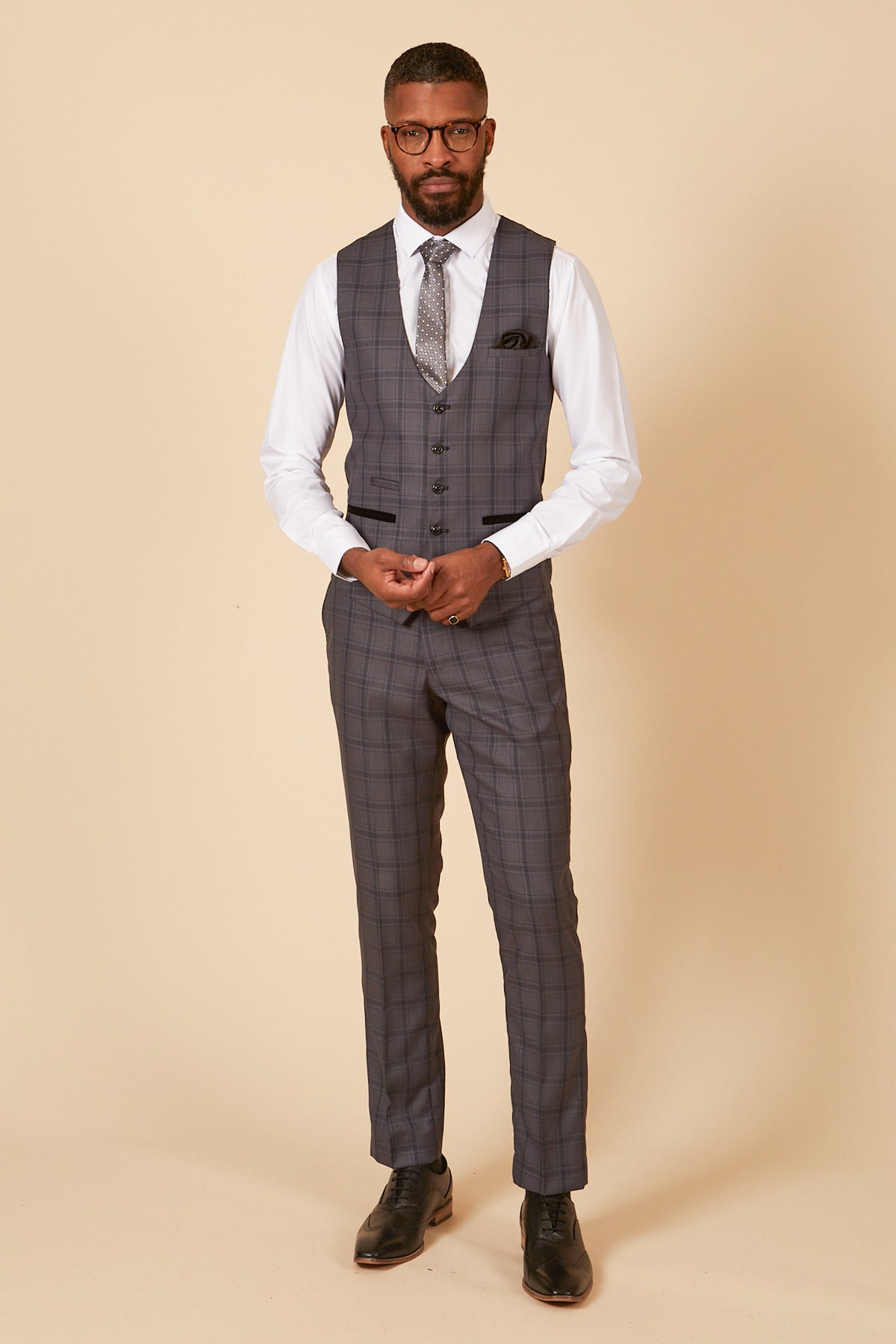 A Black man wearing glasses, a white shirt, the marcdarcy JOSE Grey Check Three Piece Suit, a patterned tie, and black dress shoes stands on a plain beige background.