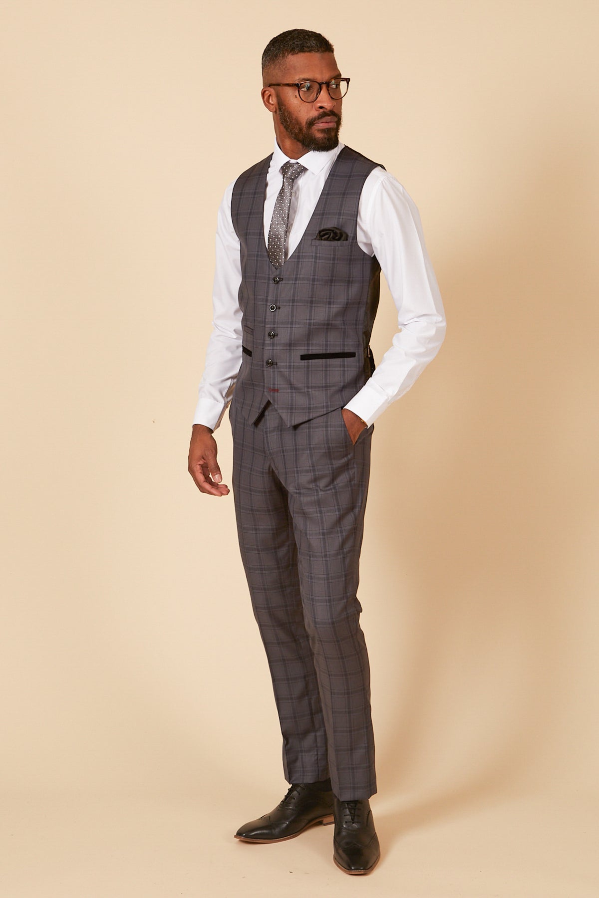 A man wears the marcdarcy JOSE Grey Check Three Piece Suit—glasses, white shirt, patterned tie, and matching vest and pants—standing against a beige background, looking to the side with one hand in his pocket.