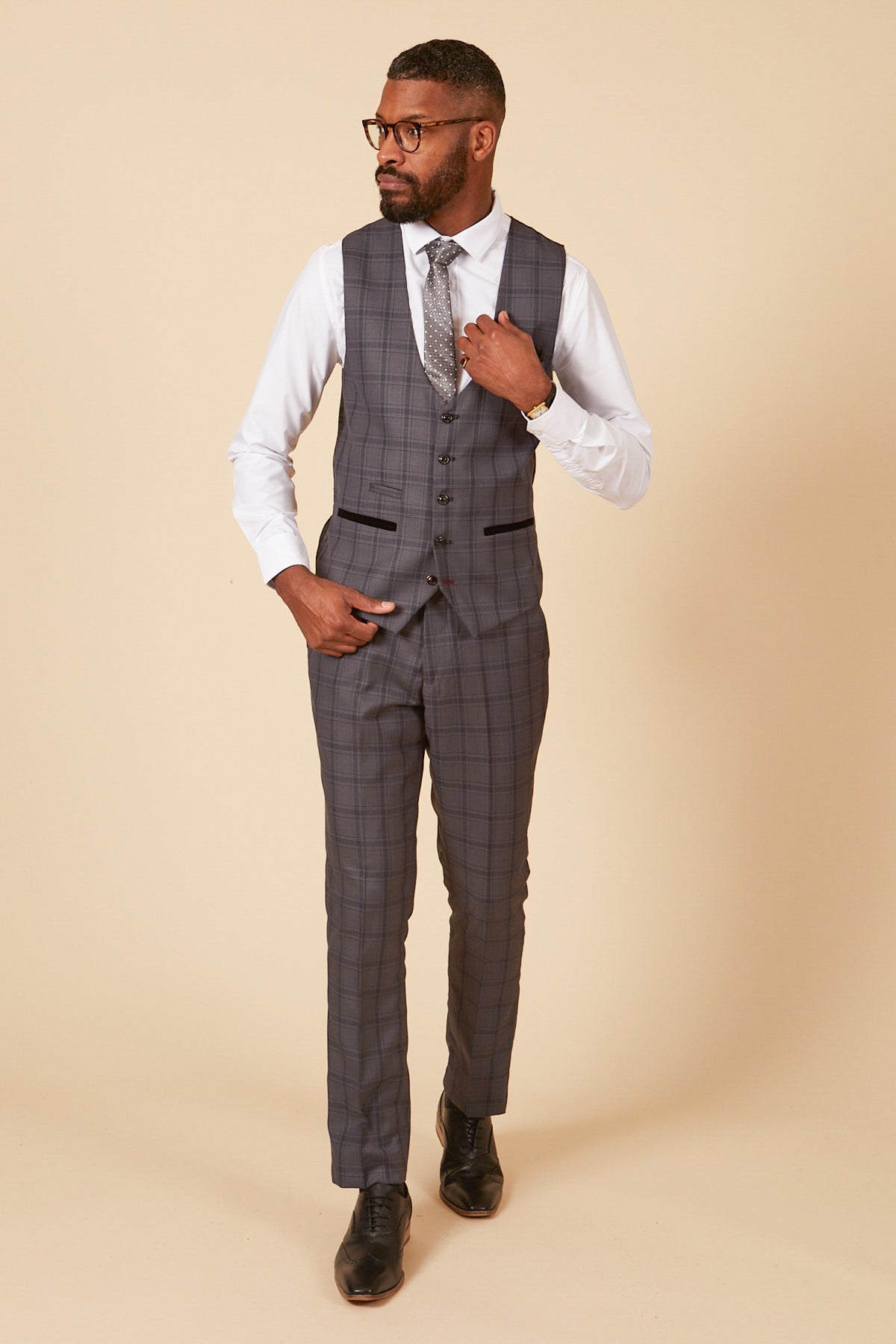 A man wearing glasses poses in a marcdarcy JOSE Grey Check Single Breasted Waistcoat, white shirt, patterned tie, and black shoes against a beige background. He adjusts his vest with one hand and looks to the side.